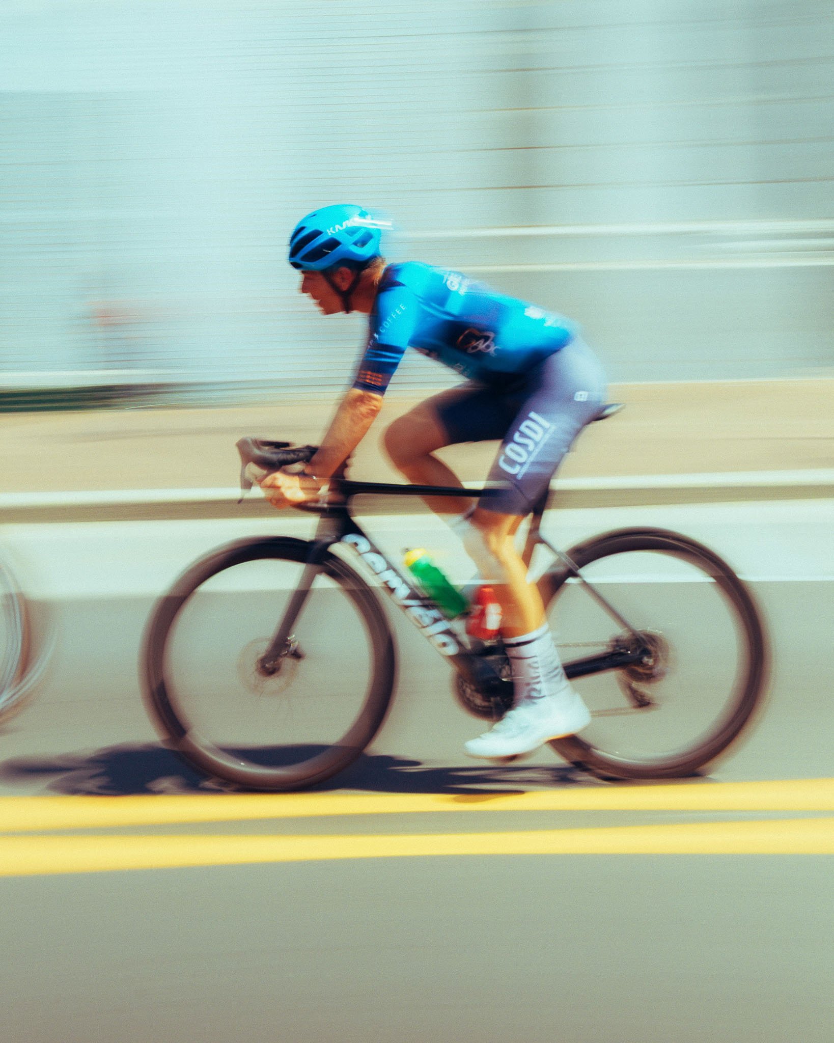 A cyclist wearing a blue helmet and blue jersey riding a black road bike on a smooth, light-colored surface with horizontal lines in the background.