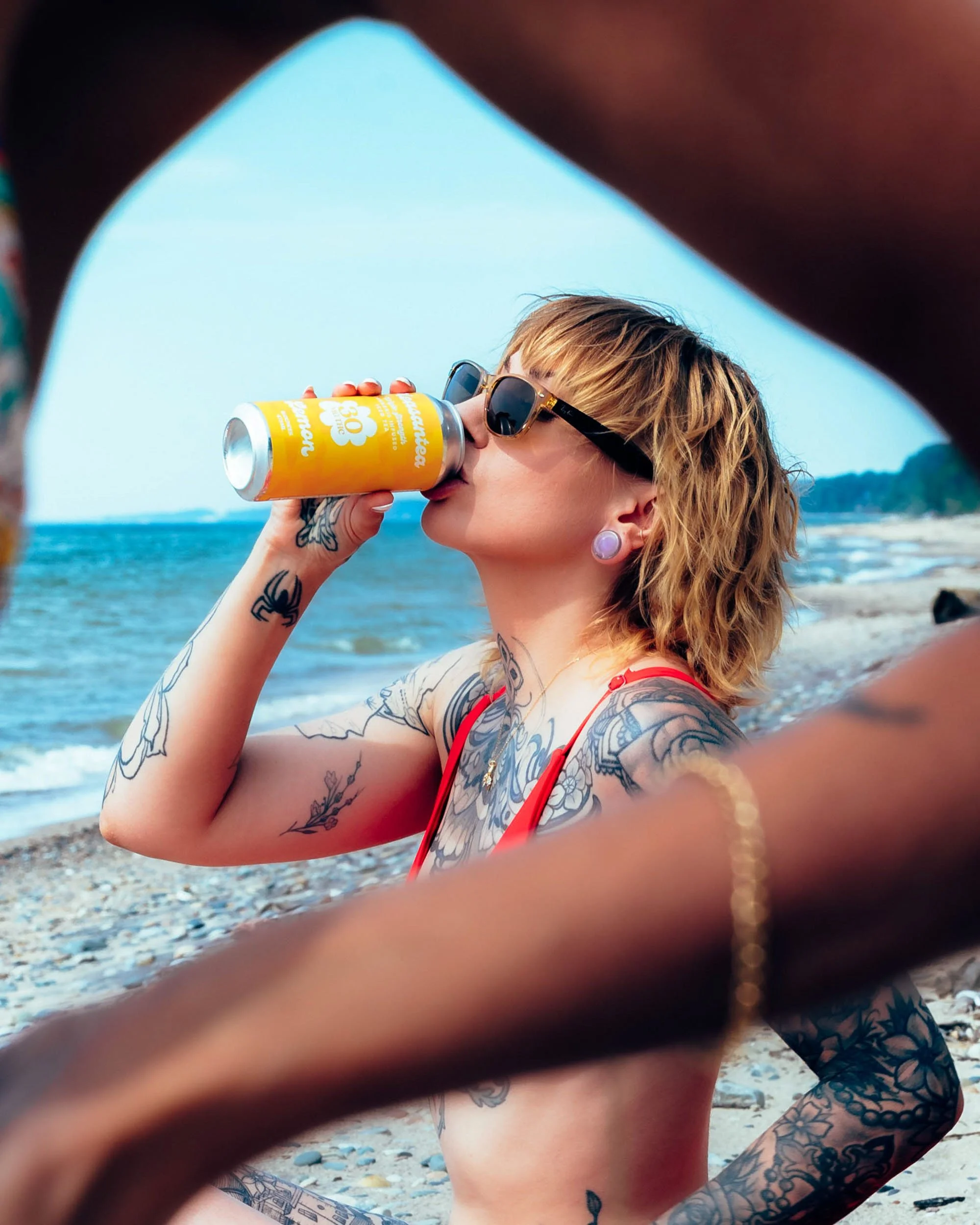 Woman with tattoos and sunglasses drinking from a yellow can at the beach, seen through a frame created by arms and fingers.