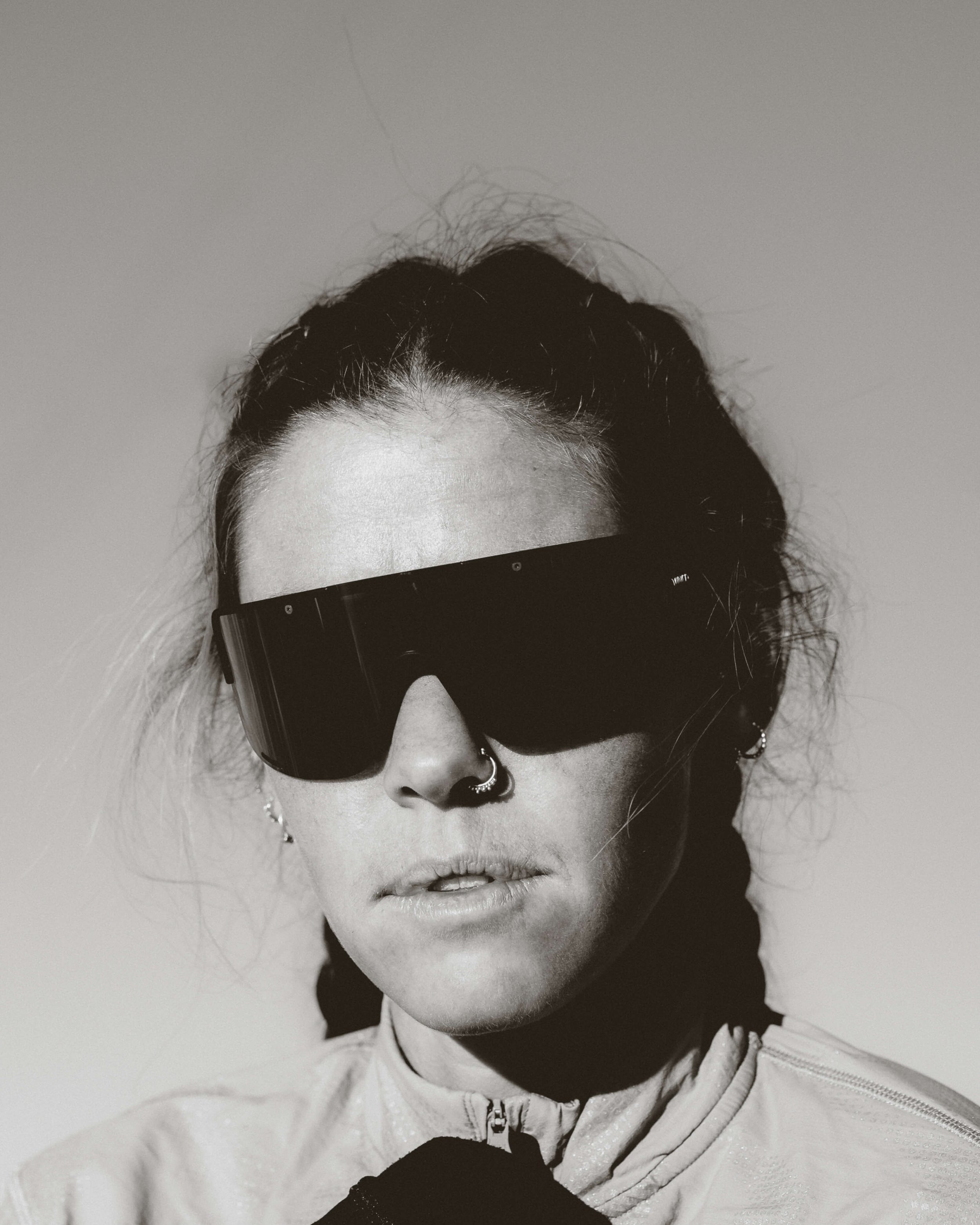 A black and white photo of a woman with dark hair, wearing sunglasses, a nose ring, and earrings, and dressed in a hoodie.
