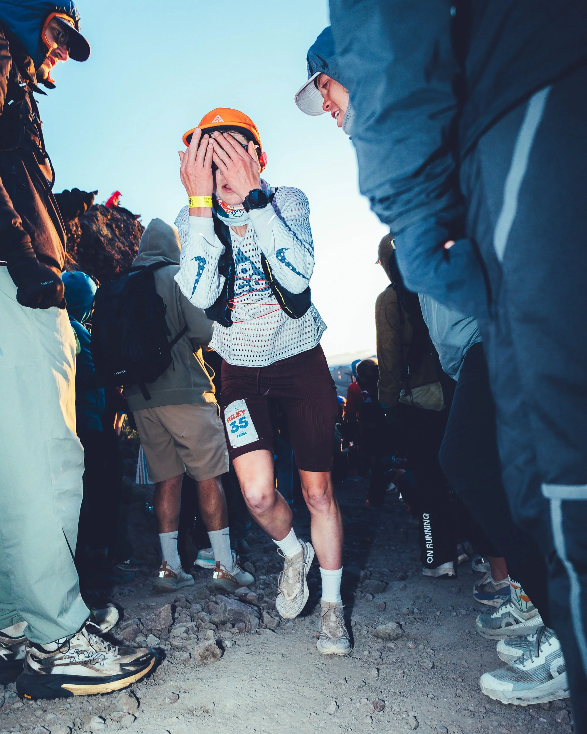A group of runners in athletic gear, gathered outdoors on a rocky dirt trail, with one woman in the center covering her face with her hands, wearing a white long-sleeve shirt, maroon shorts, and a bright orange hat, surrounded by others in jackets, h