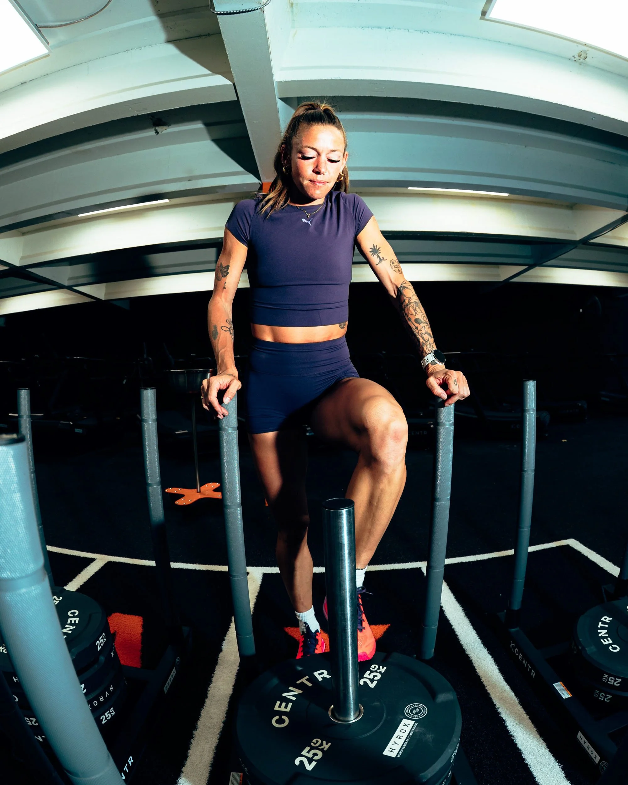 A woman with tattoos on her arms wearing a purple sports shirt and shorts, exercising at a gym. She is stepping over a weight plate on a workout machine, with her eyes closed trying to focus. The gym has multiple weight plates labeled '25 KG' around 