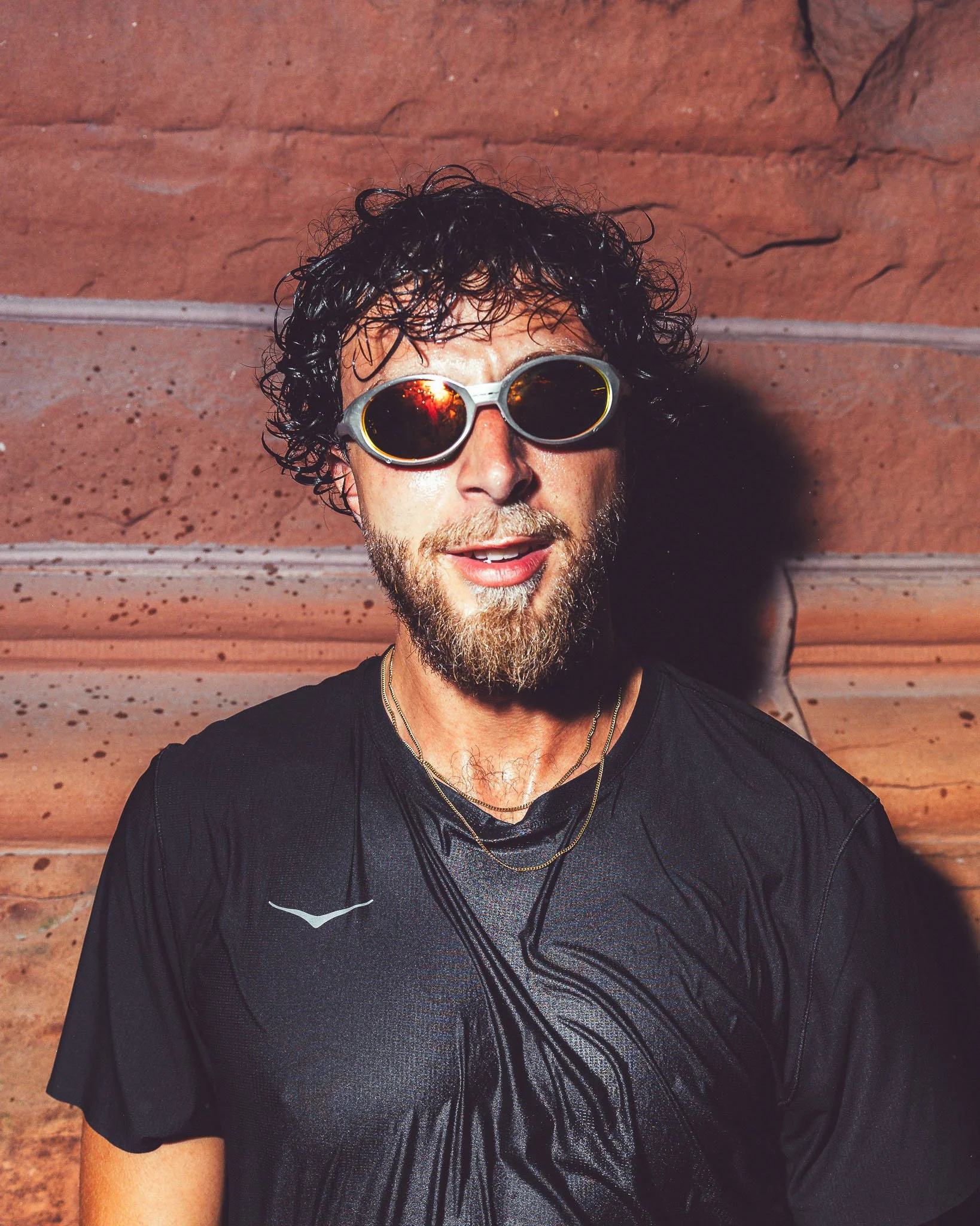 A man with curly dark hair, a beard, and sunglasses standing against a textured reddish-brown background, wearing a black shirt with a shiny black design on the front.