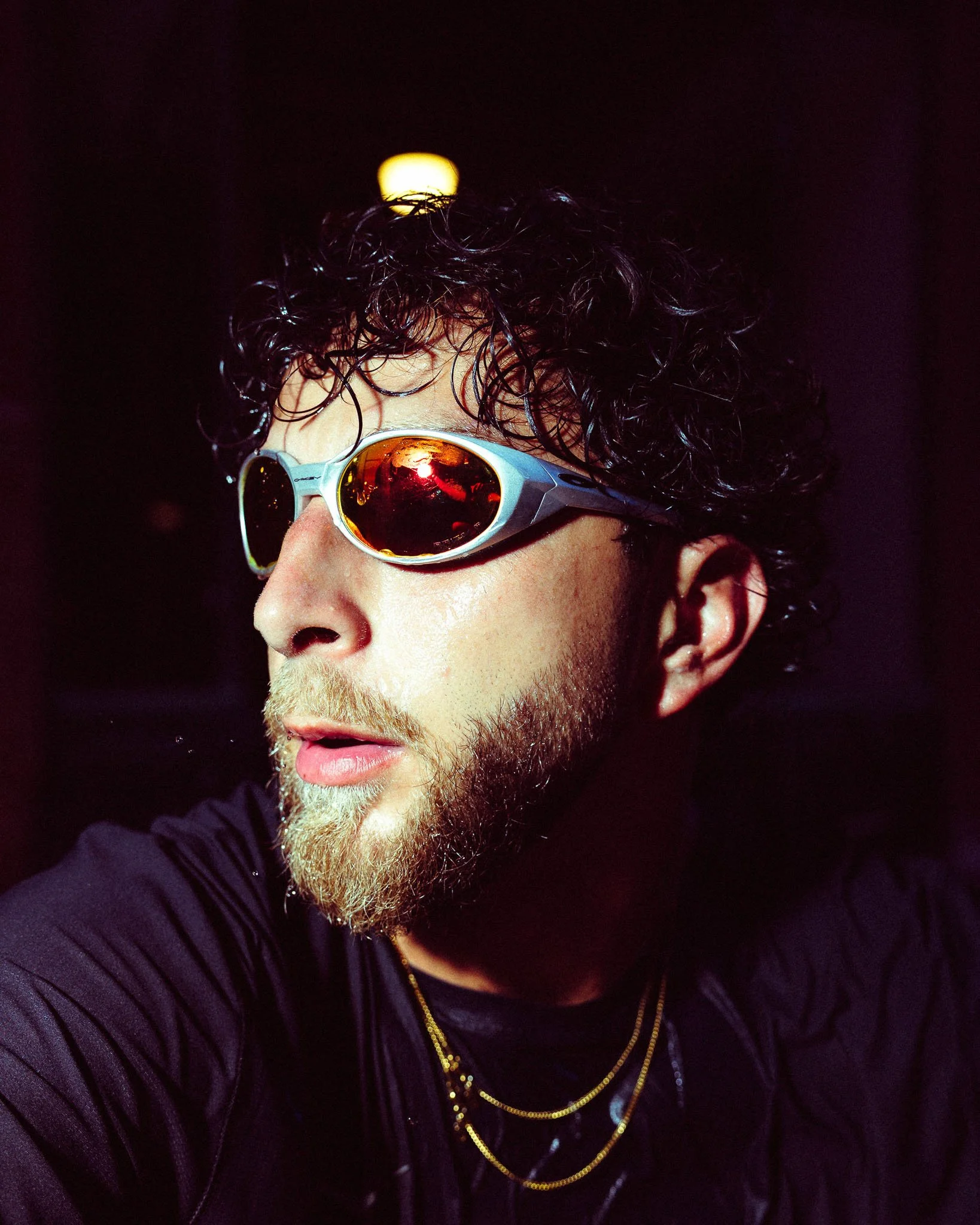 Close-up of a man wearing sunglasses with reflective orange lenses, curly hair, and a beard, against a dark background with a bright light source behind his head.