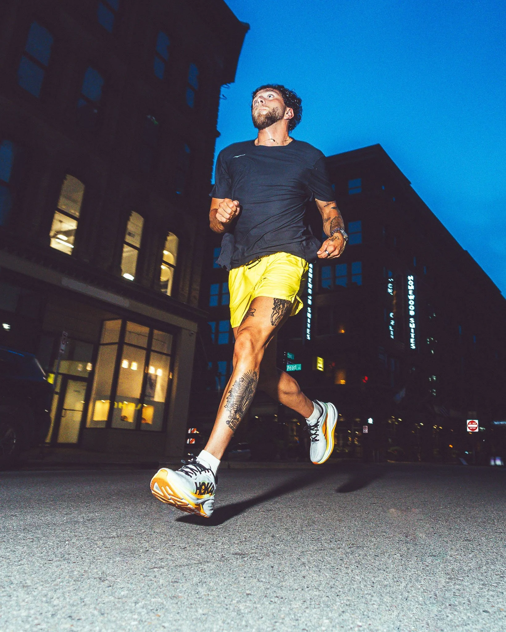A man running on a city street at night, wearing a black t-shirt, yellow shorts, and white Nike sneakers, with tattoos on his left leg and right arm, as buildings with lit windows and signs are visible in the background.