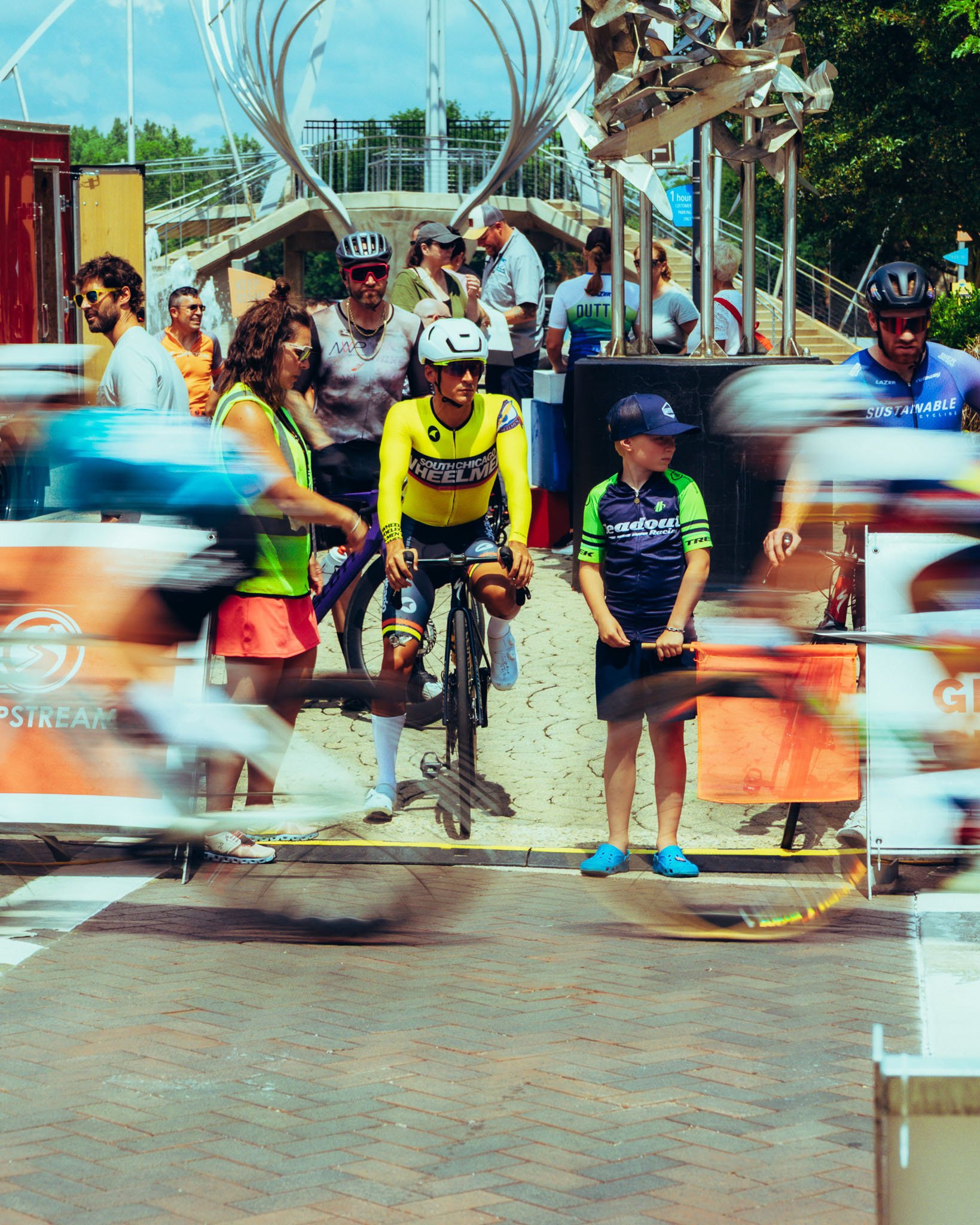 Group of cyclists and event staff near start line at outdoor cycling race, with some blurred motion from passing bikes, in bright sunny weather.