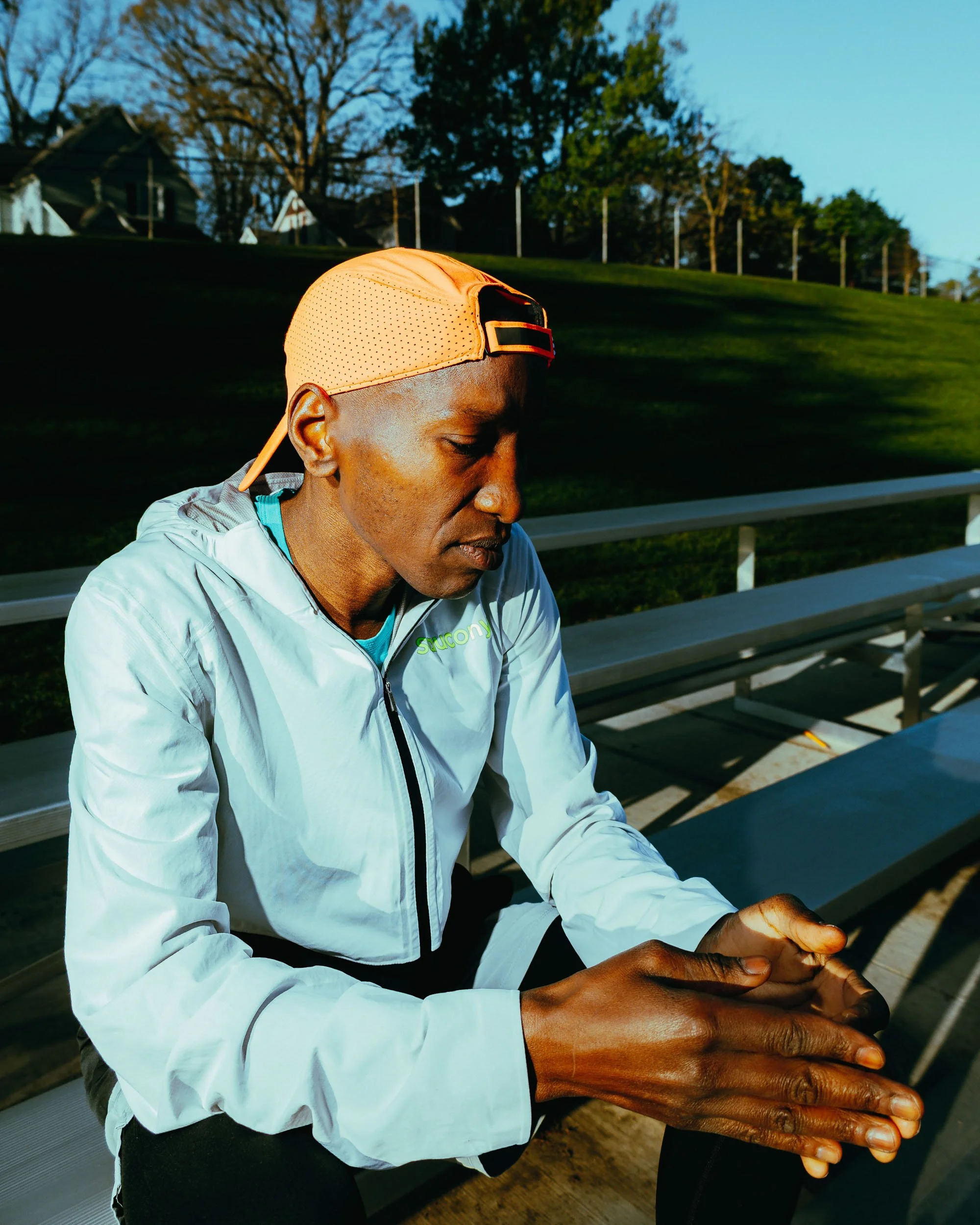 A man with dark skin, wearing a white athletic jacket and a light orange cap, is sitting on a park bench outdoors with his eyes closed and hands clasped, possibly praying or meditating.