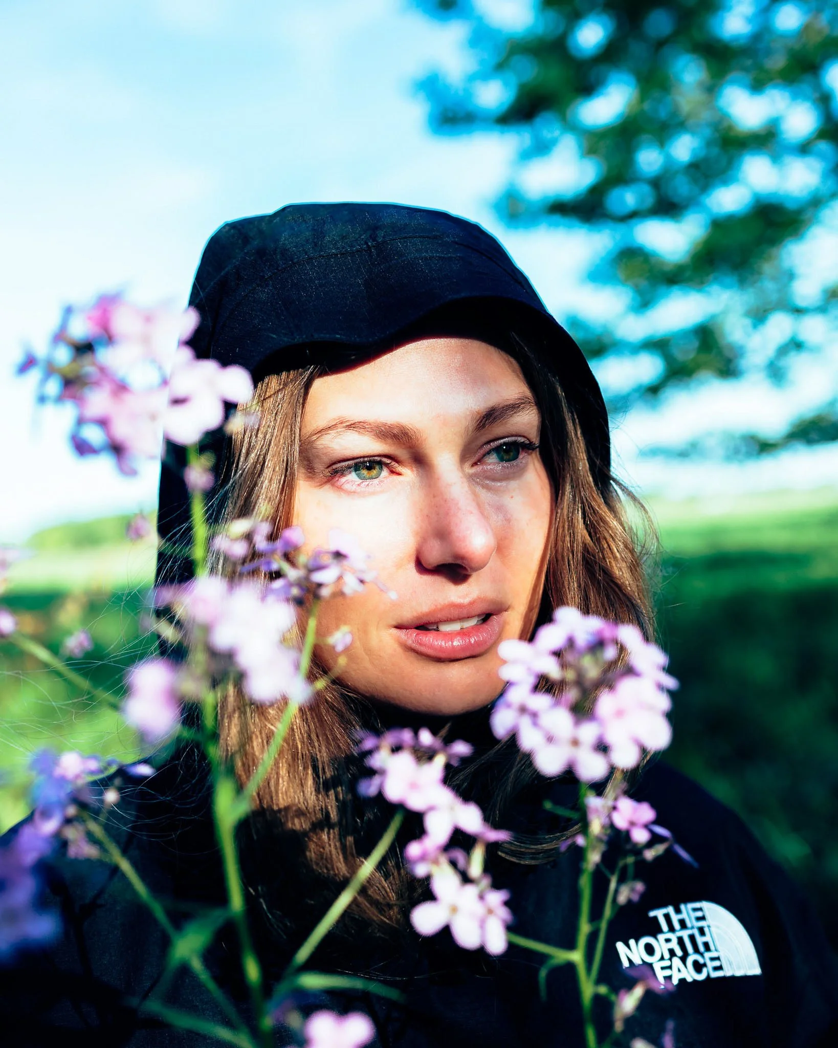 Close-up of a woman with long brown hair and green eyes, wearing a black cap and black shirt with The North Face logo, partially obscured by pink flowers outdoors with green trees and blue sky in the background.