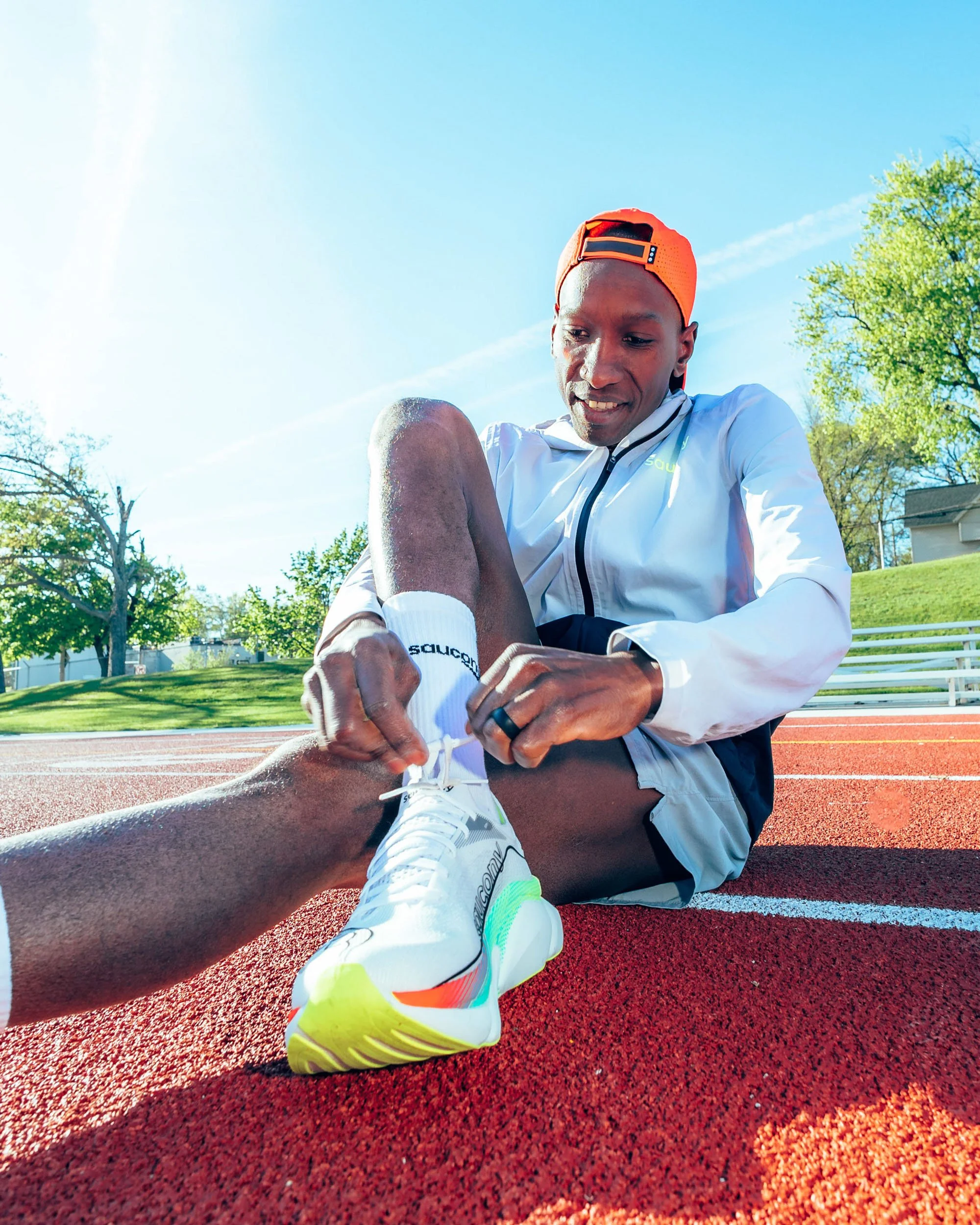 A runner sitting on a red track field outdoors, tying his shoe with another person helping. The runner is wearing a white athletic jacket, neon yellow and orange running shoes, white socks, and an orange cap worn backwards. The background features tr