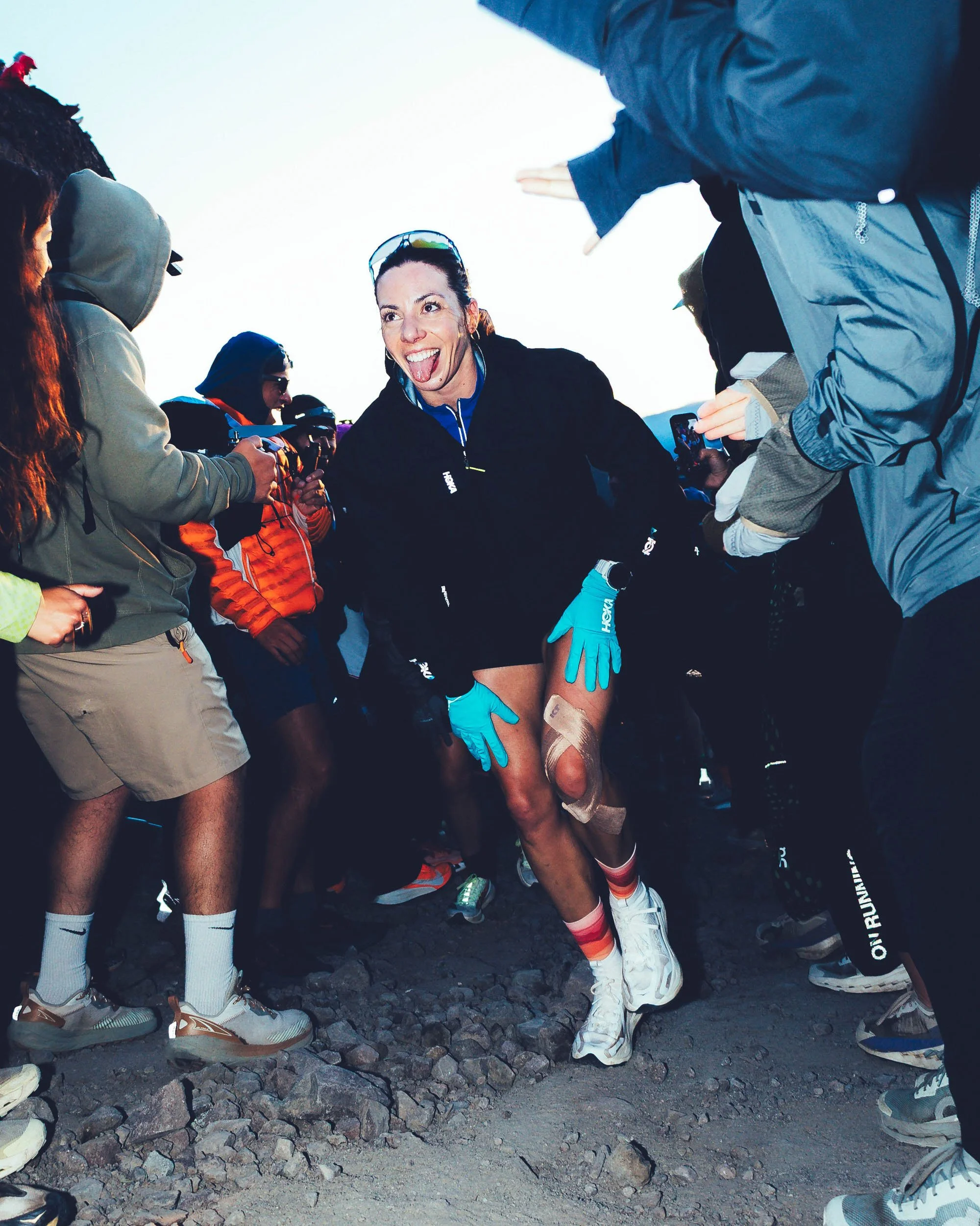 A group of people standing outdoors on rocky ground, with one woman in the center smiling and sticking her tongue out, wearing a black jacket, gloves, and athletic gear, surrounded by others dressed in outdoor clothing.