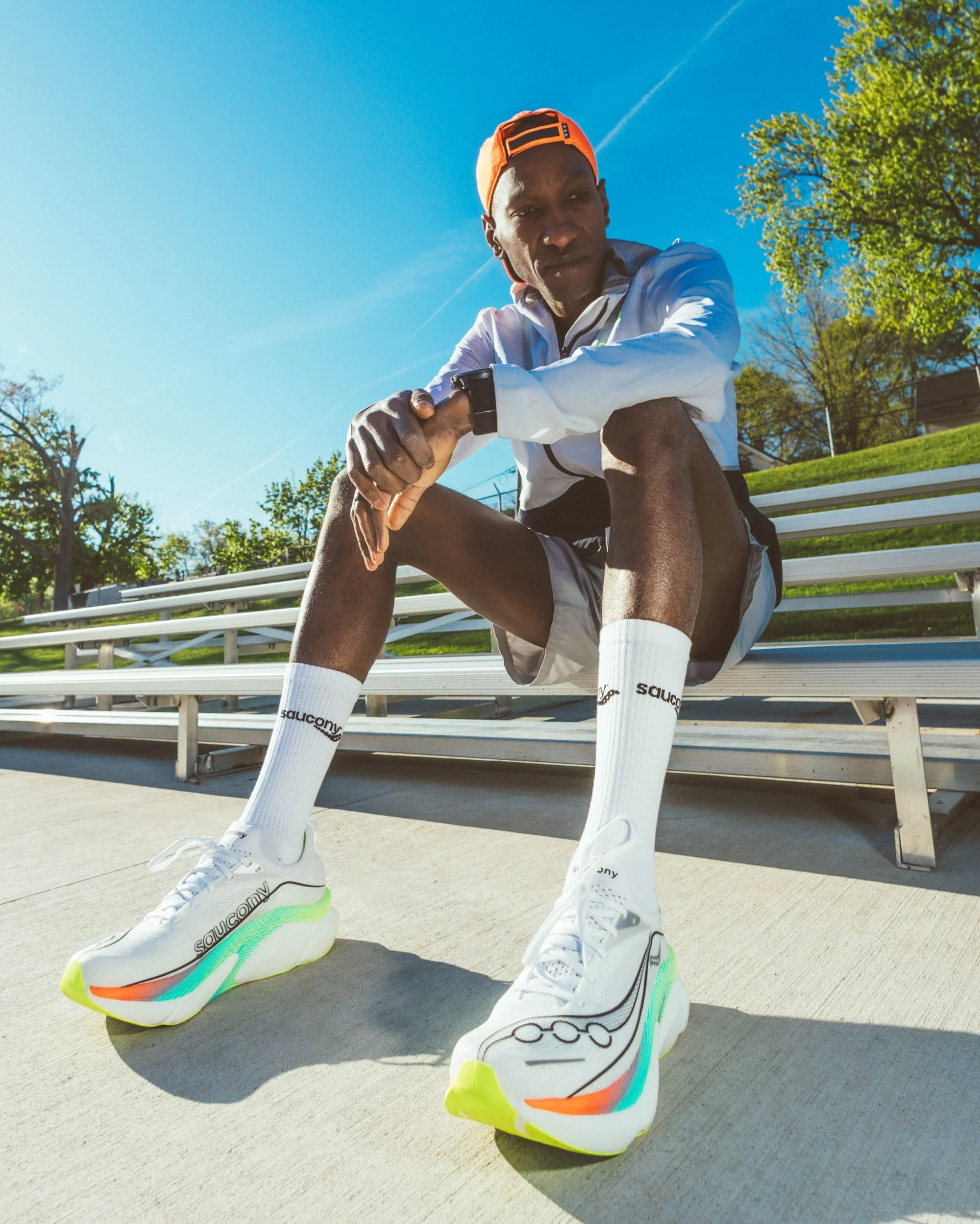 A man in sportswear sitting on a bench outdoors, wearing white sneakers with colorful accents, white socks with black text, a white jacket, and an orange cap backwards, under a bright blue sky, with trees and a grassy area in the background.