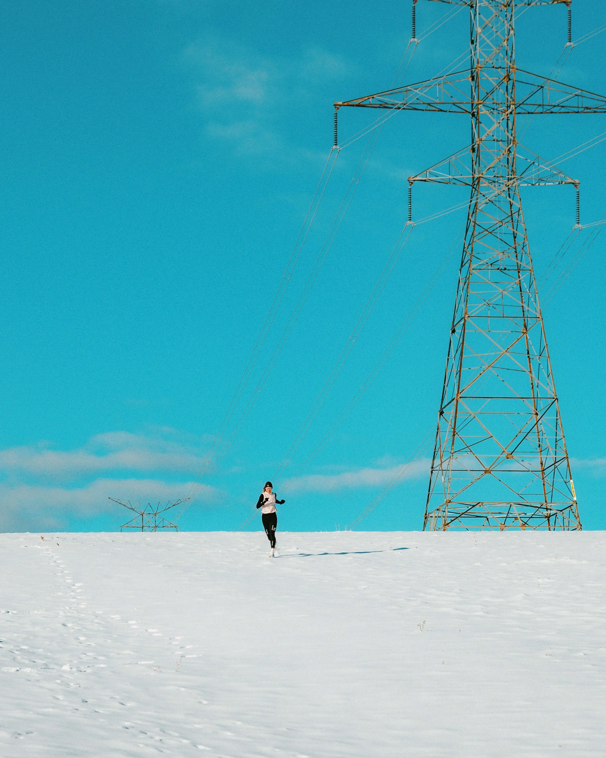 A person jogging on snow-covered ground with power lines and electric towers in the background on a clear, blue sky day.