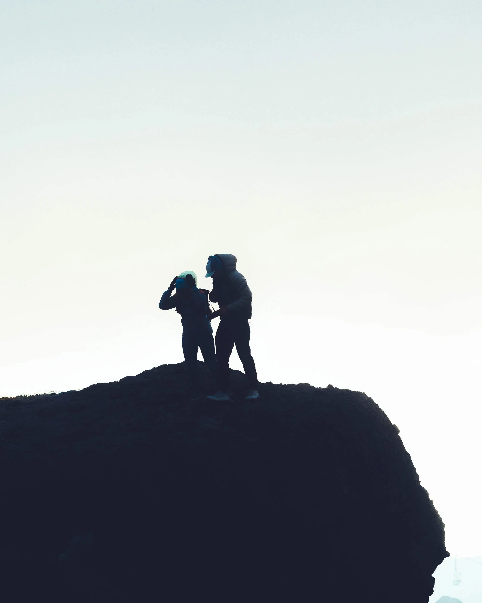 Two people wearing winter gear standing on a rocky mountaintop, looking at the sky.