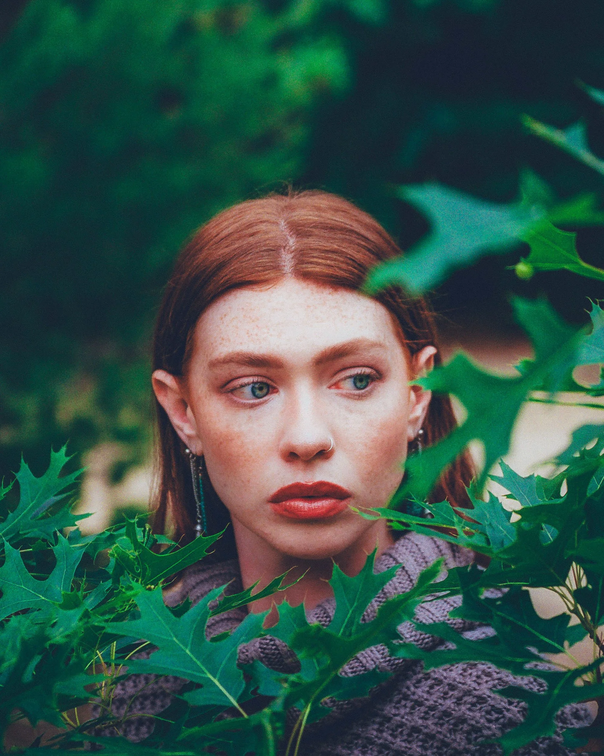 A woman with red hair and blue eyes looking through green holly leaves.