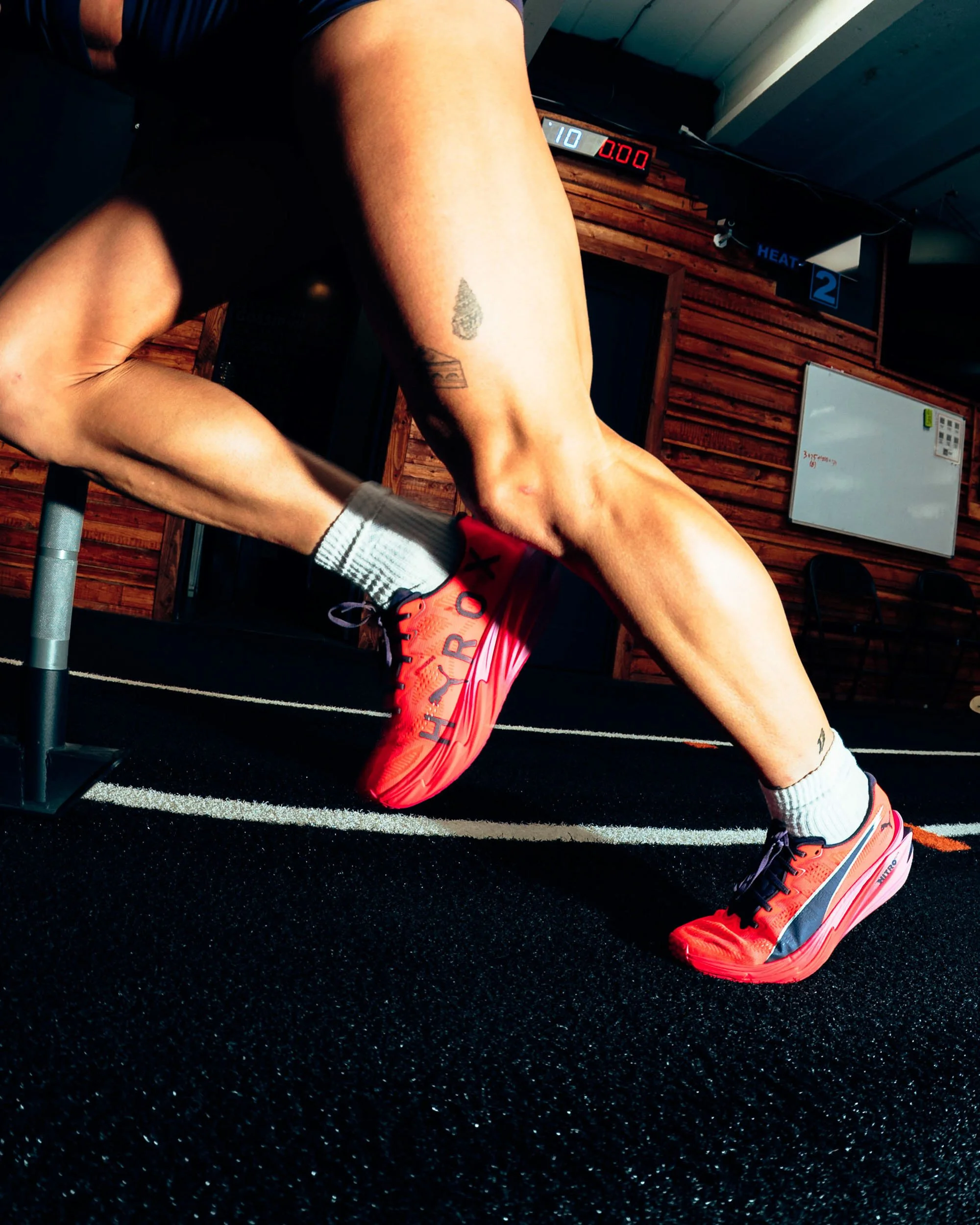 Close-up of an athlete's legs in a workout gym, with a focus on the calf and knee, wearing orange and black sneakers and gray socks, on black rubber gym flooring, with a wooden wall and a whiteboard in the background.