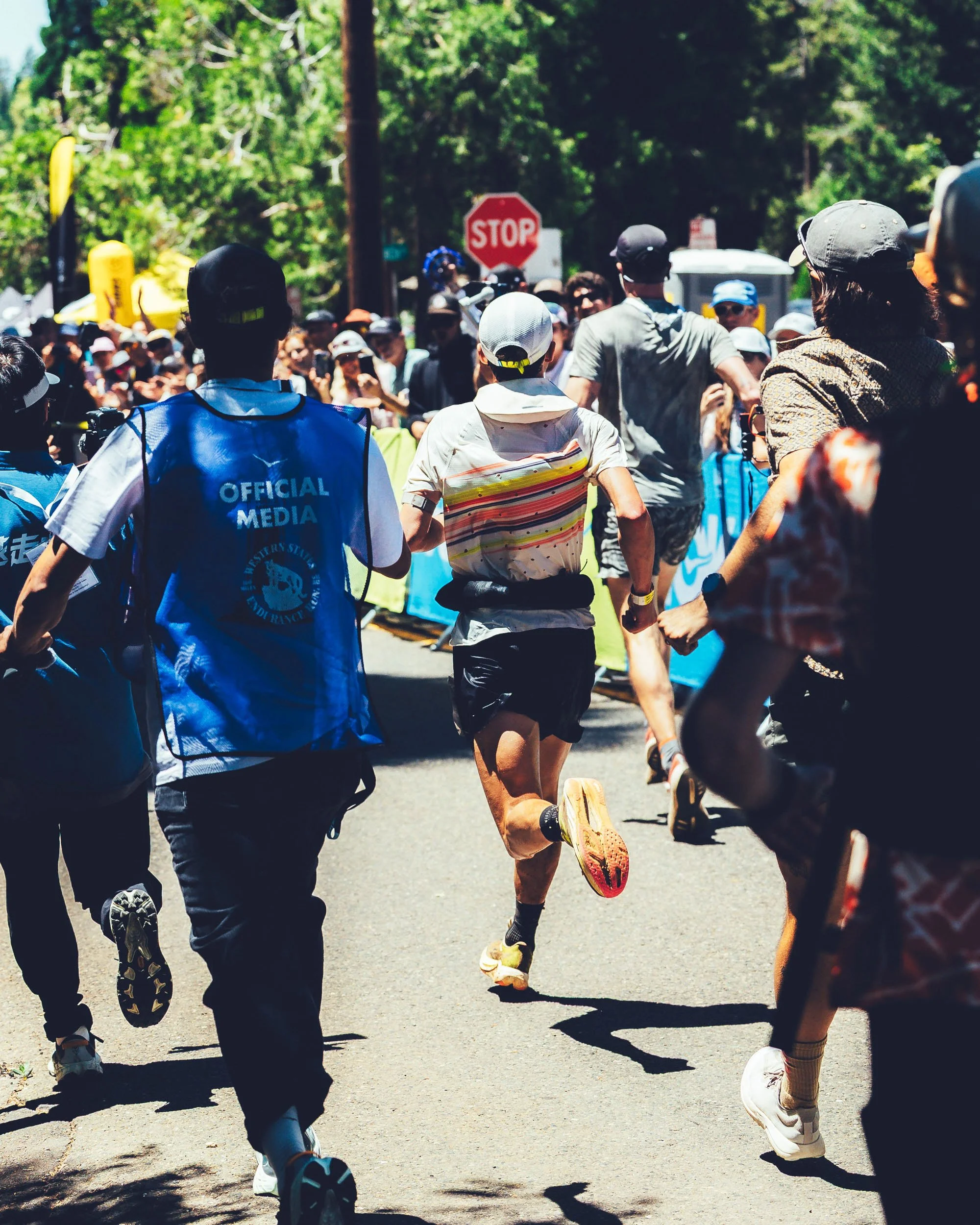 A group of runners participating in a race, running on a tree-lined road with a stop sign in the background.
