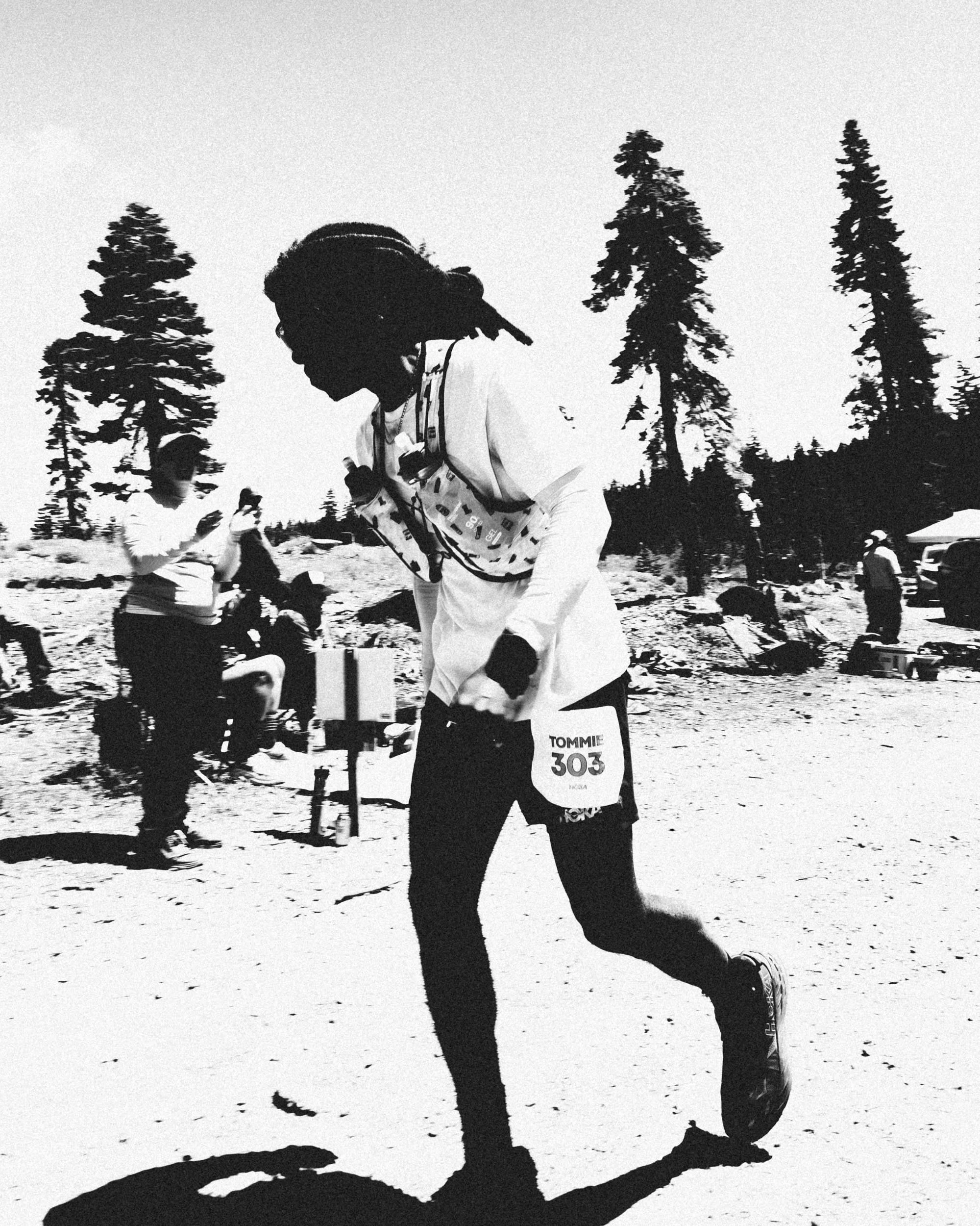 A person wearing athletic gear and a bib with the number 303, participating in an outdoor running event, with tall trees in the background and a mostly clear sky.