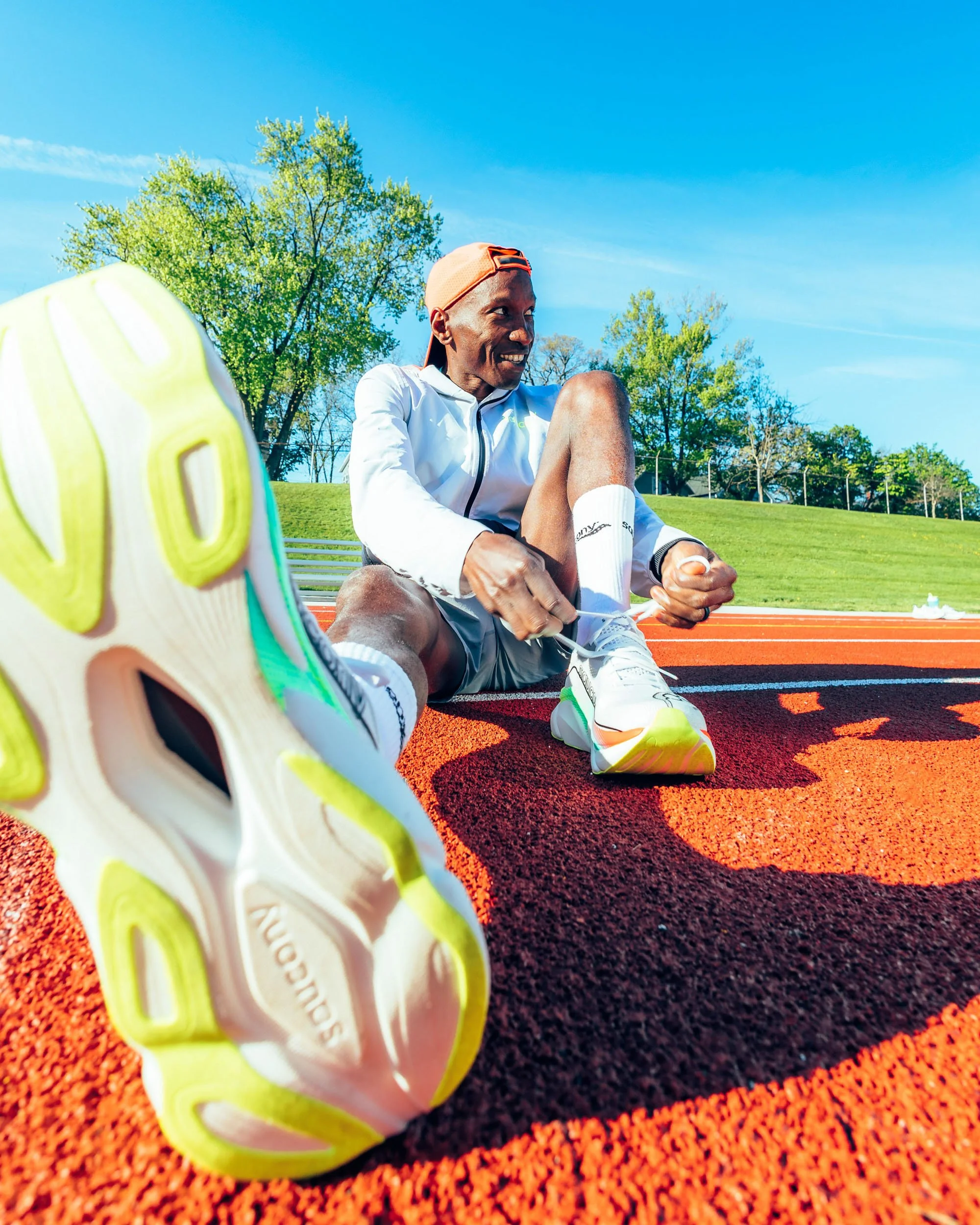 A man sitting on a running track tying his shoelaces, with a focus on his running shoe in the foreground. The man is wearing athletic clothes, including a white jacket, white shorts, and a beige cap. The background features trees and a bright blue sk
