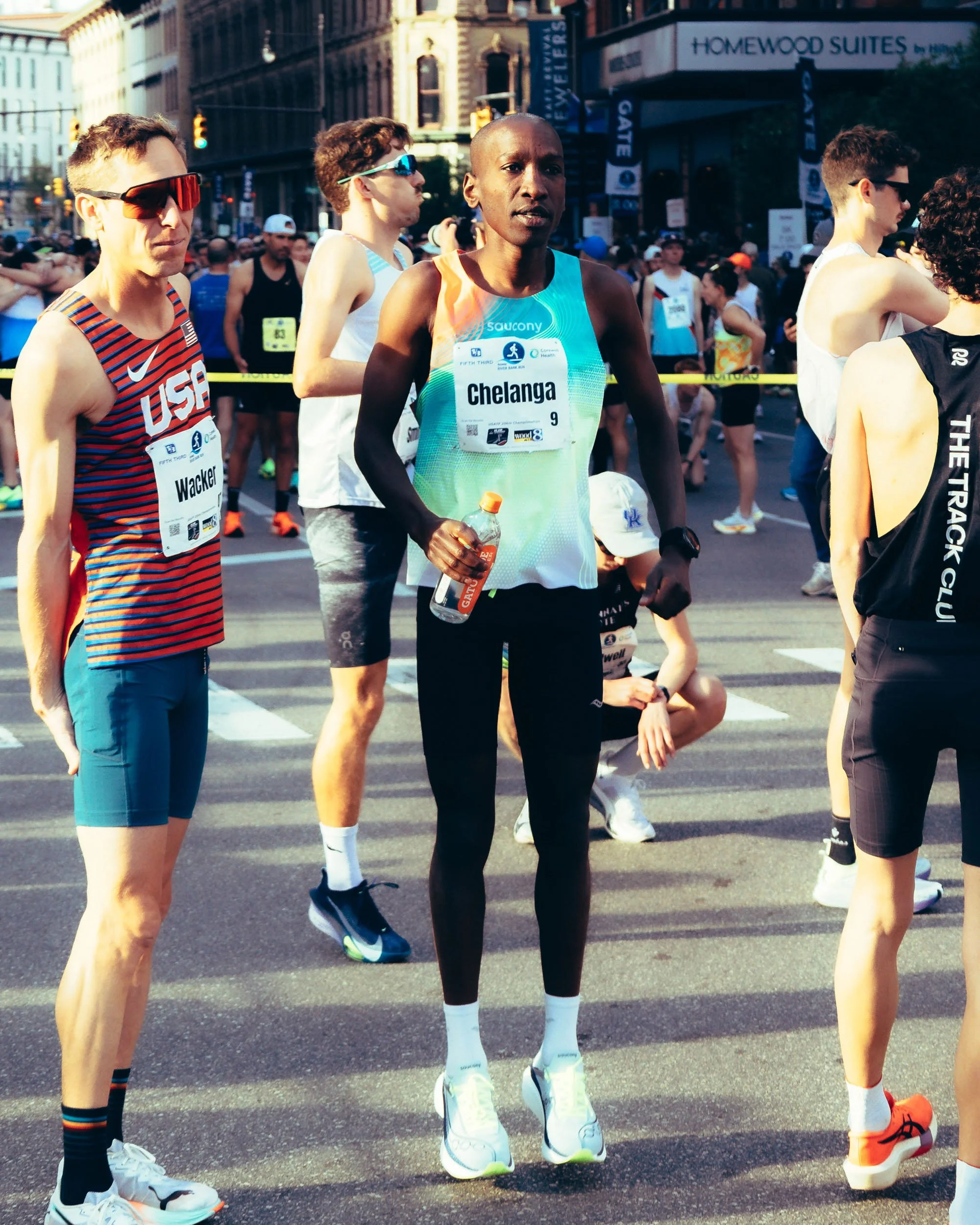 Group of marathon runners preparing at the start line in an urban setting during daytime. A woman in the center wears a colorful tank top with the name Chelanga. Runners have bib numbers and athletic gear, with some wearing sunglasses and others hold