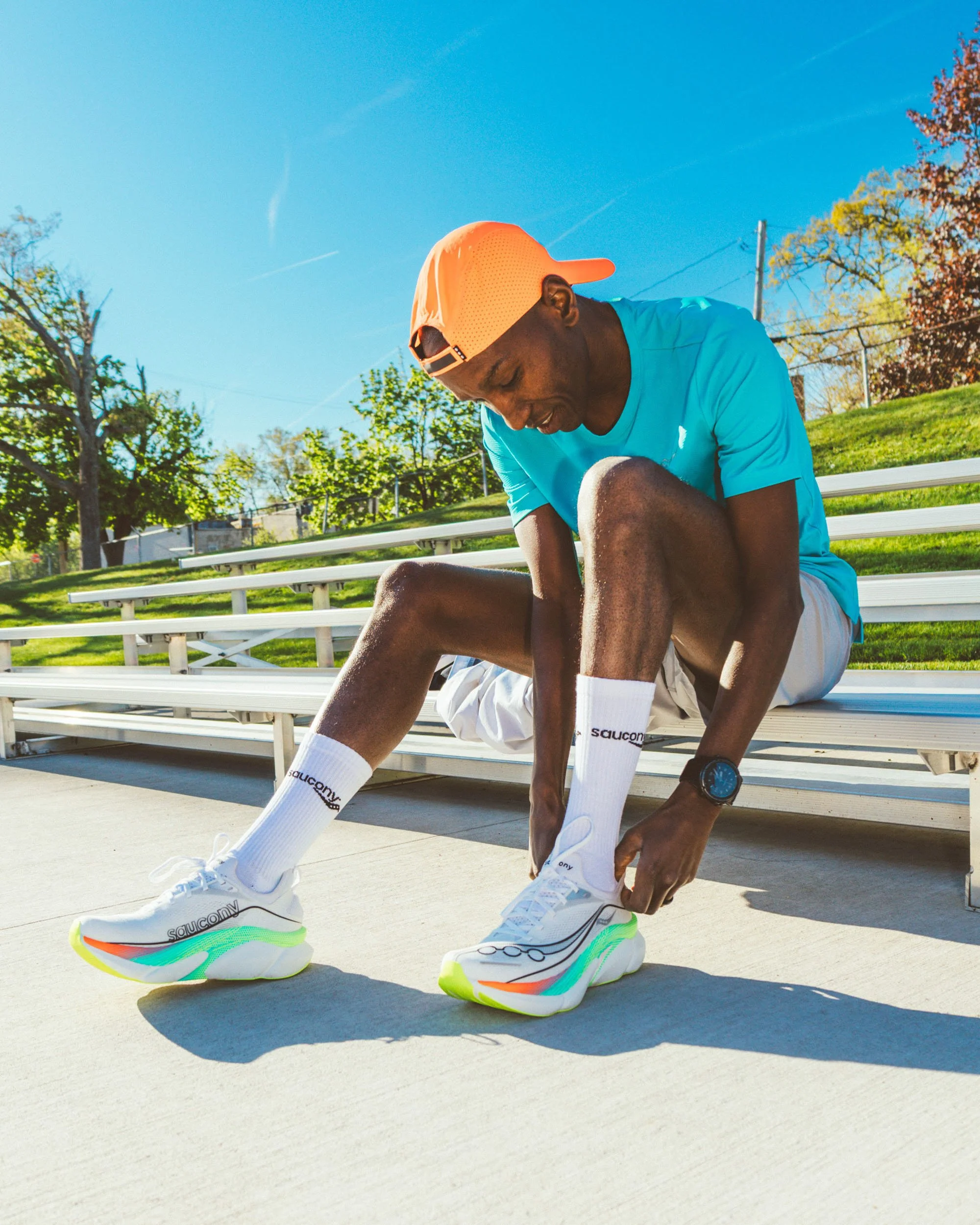 Two men sit on bleachers outside on a sunny day, with trees and a fence in the background. One man is adjusting his shoe while the other man is smiling and leaning in close to him, both wearing athletic clothing and sneakers.