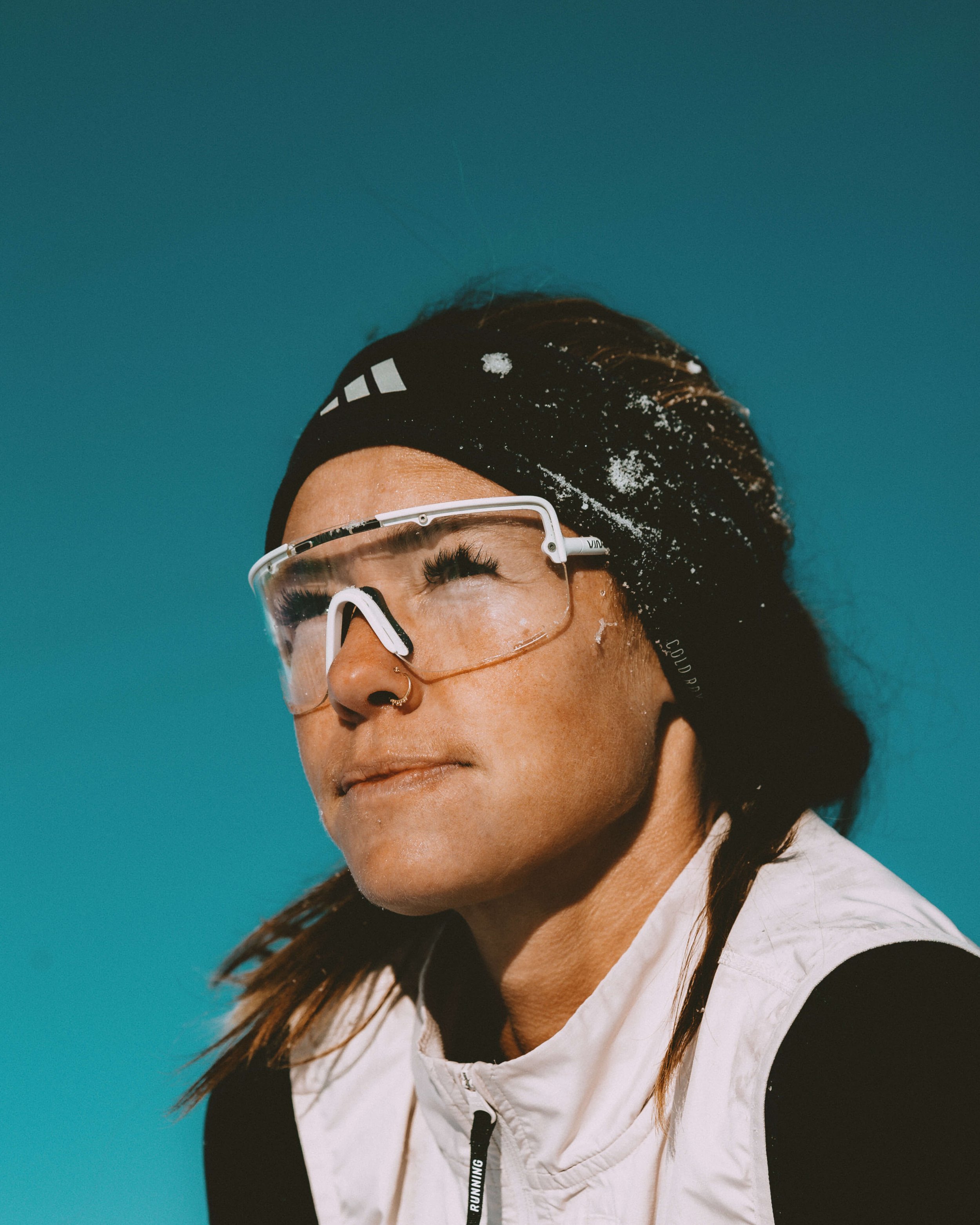 Close-up of a person with long dark hair wearing glasses, a black headband with white logo, black and white sports clothing, and a white jacket, against a teal background, with snow on their hair and face.