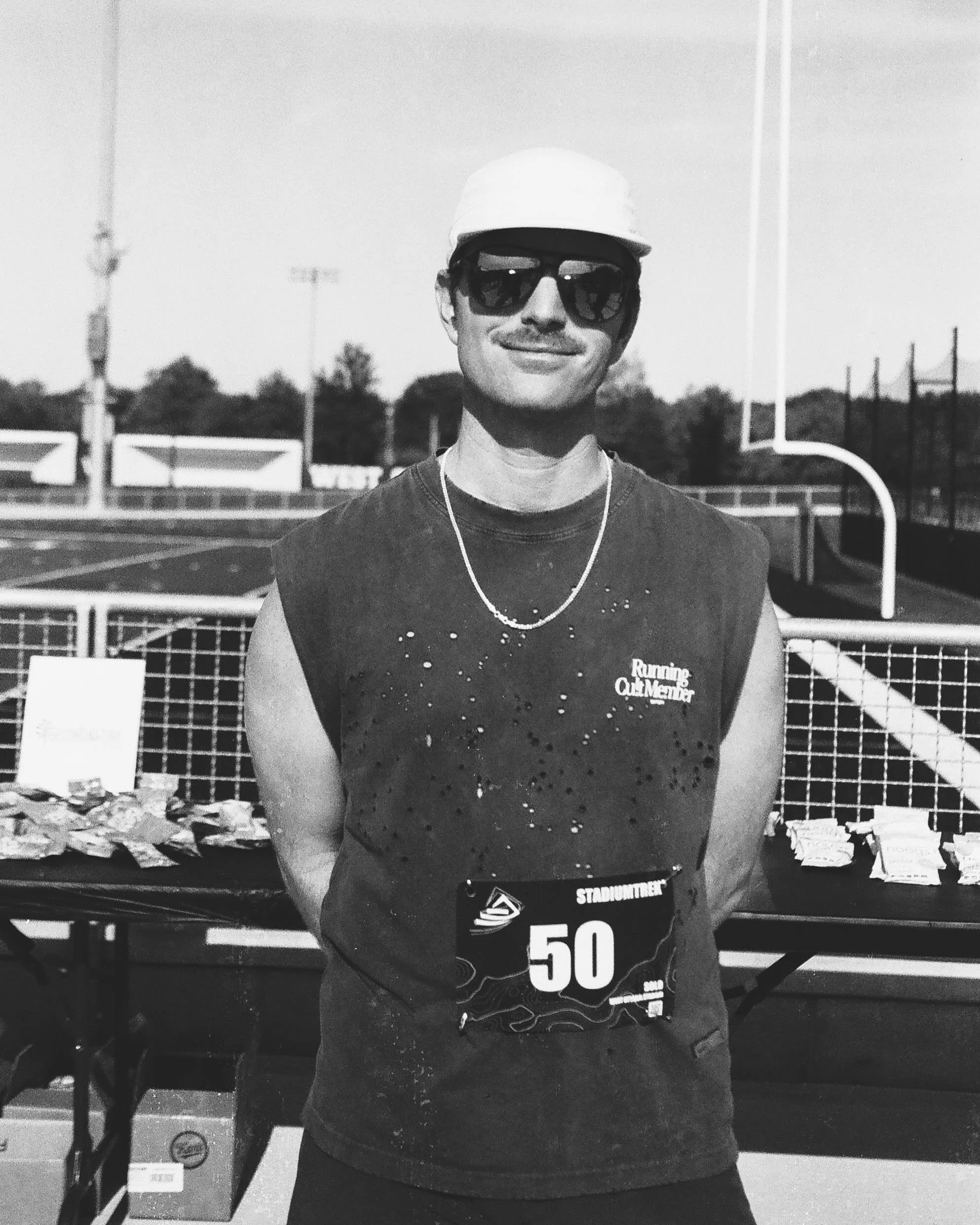A smiling man wearing sunglasses, a white cap, a necklace, and a running bib with the number 50, standing outdoors near a sports field.