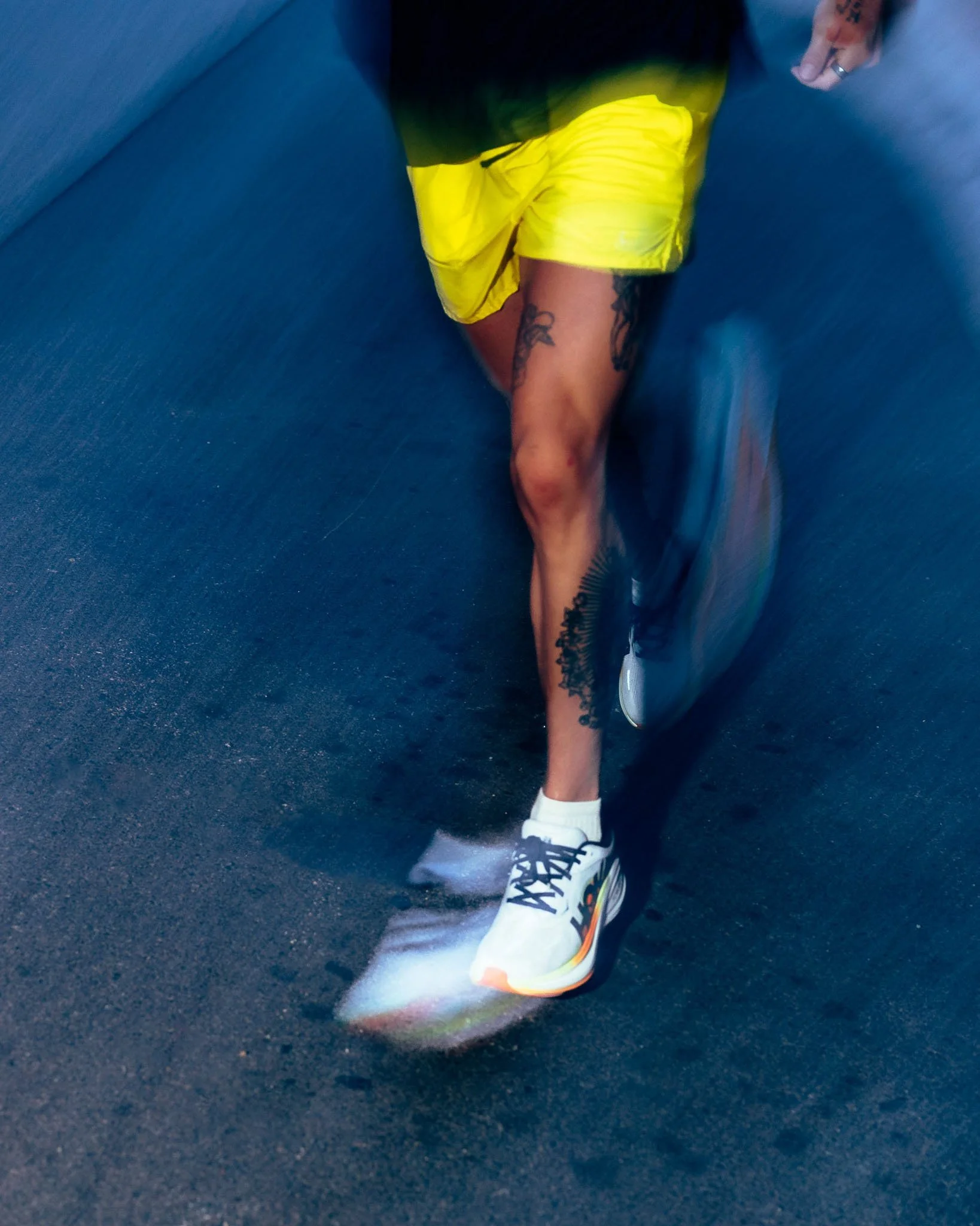Person wearing yellow shorts, white sneakers, and black socks, sitting on a blue surface, with tattoos on leg, and a rainbow-colored man bun, with part of a shoe touching the surface.