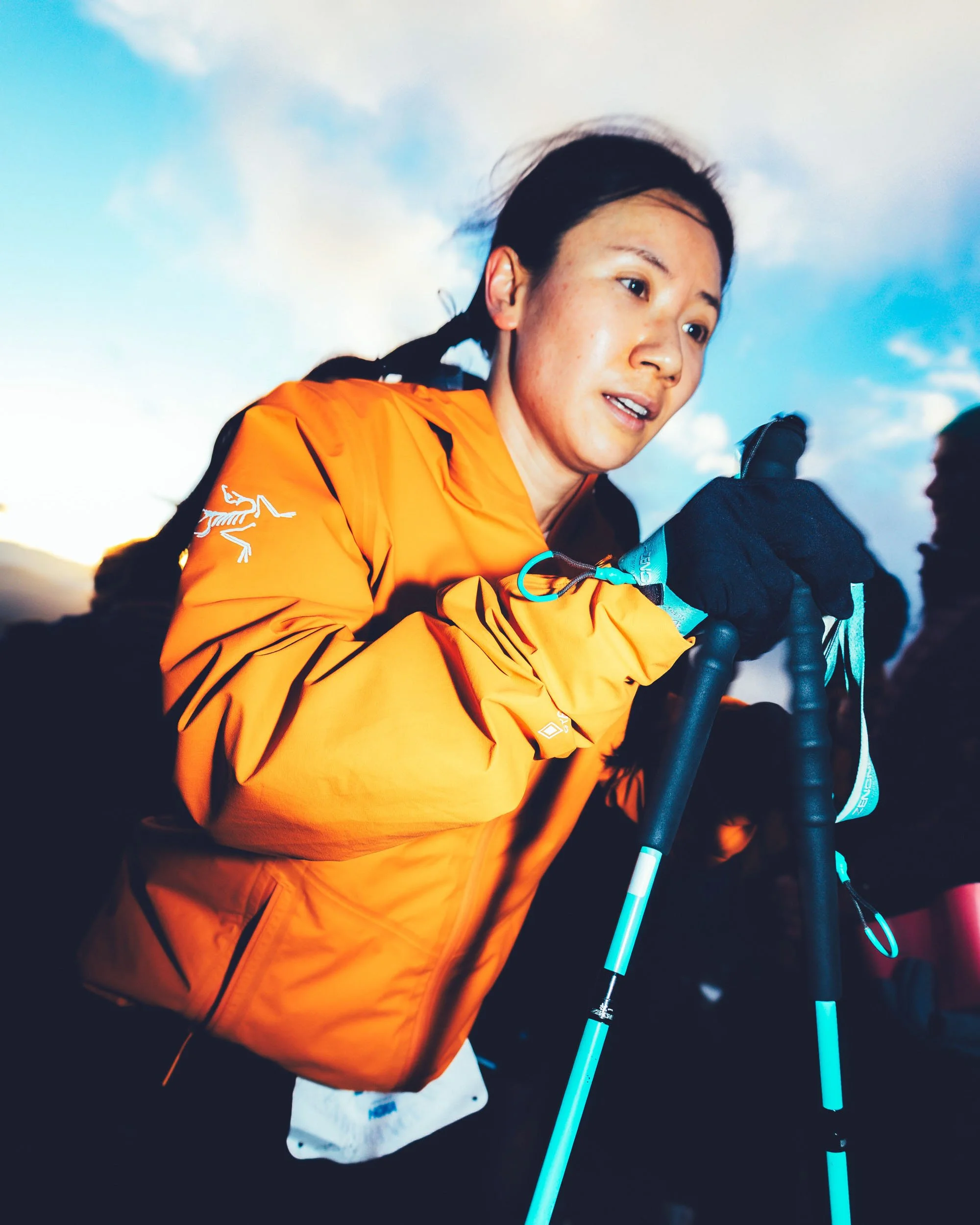 A woman with black hair tied back, wearing an orange jacket, holding ski poles, with a background of cloudy sky during sunset or sunrise.