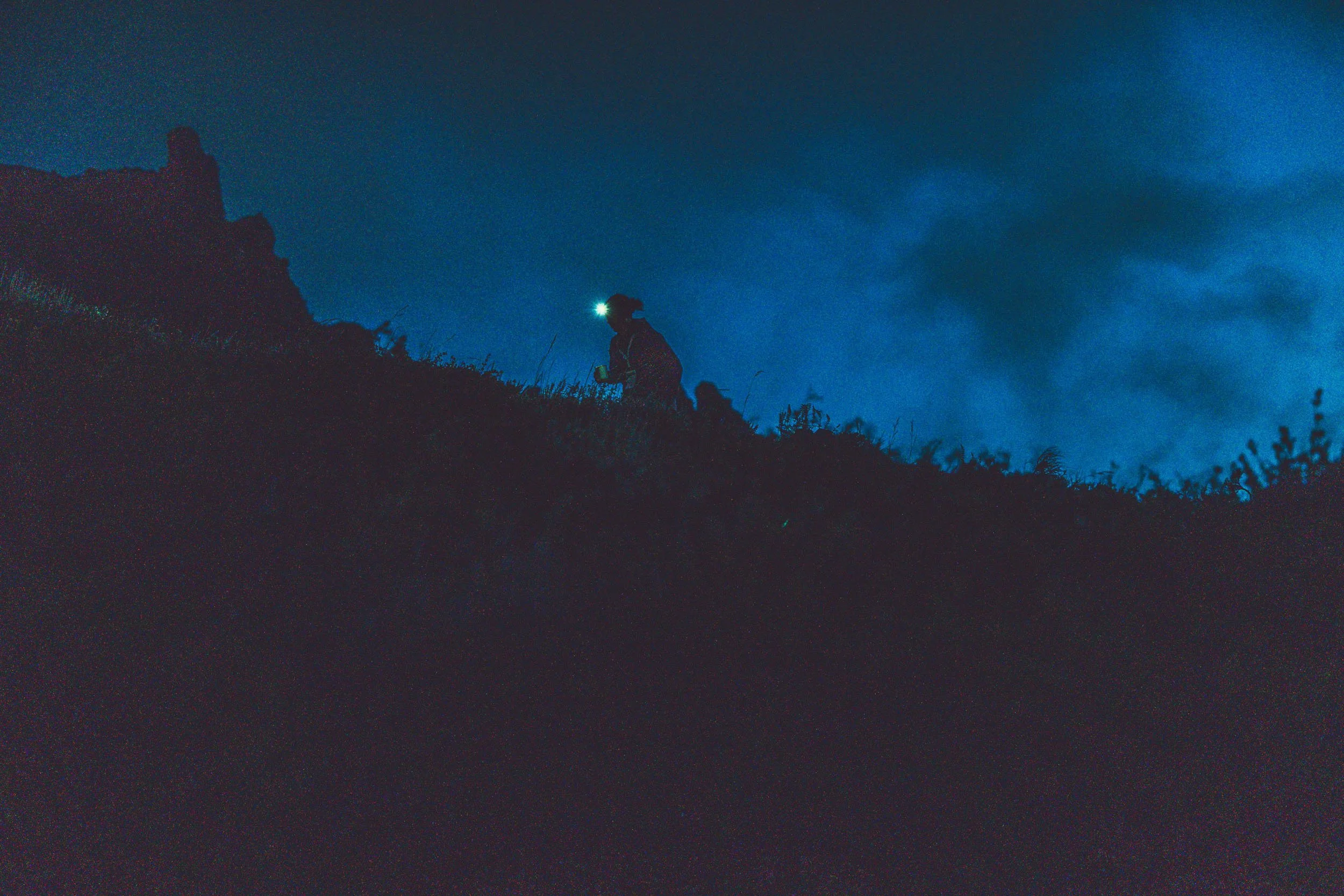 A person wearing a headlamp and outdoor clothing is trekking up a grassy hill at night, with a silhouette visible against a dark, cloudy sky.