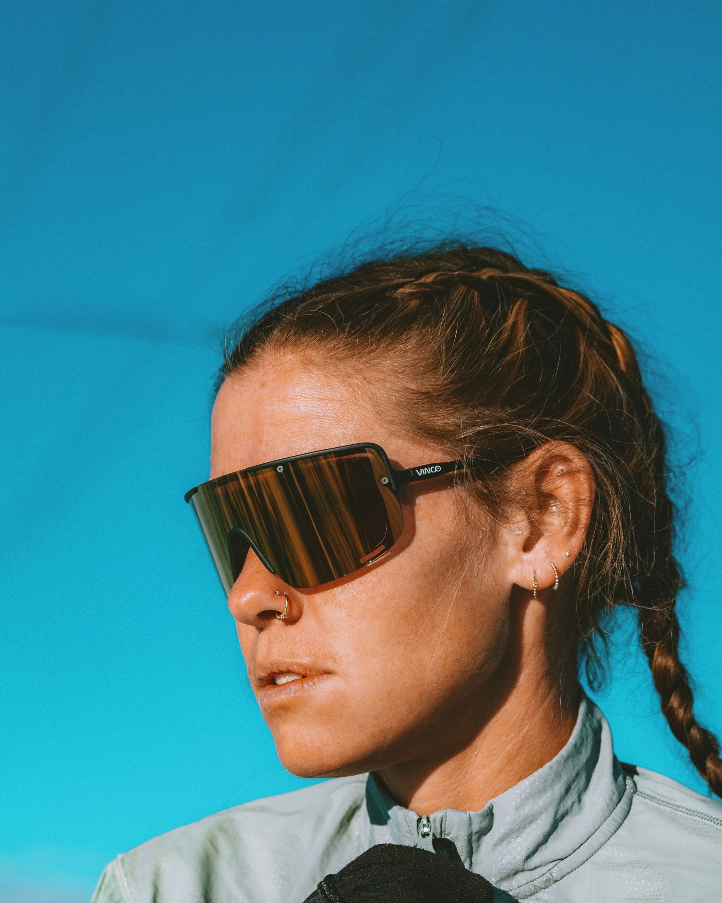 Close-up of a woman wearing large sunglasses, earrings, a nose ring, and a zip-up jacket, against a blue background.