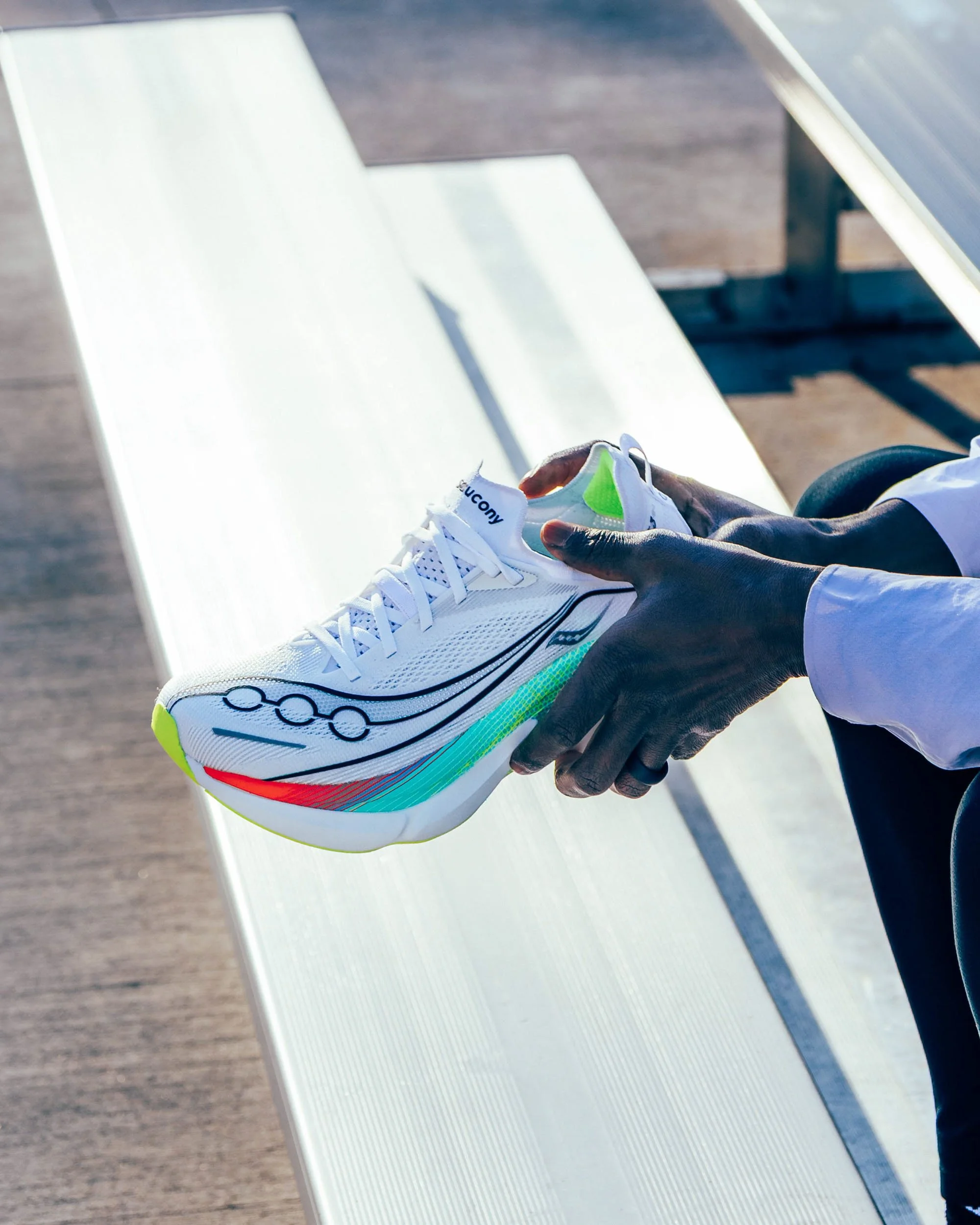 A person holding a white athletic shoe with green accents and the brand name 'Saucony' visible, in front of a white bench and wooden floor.