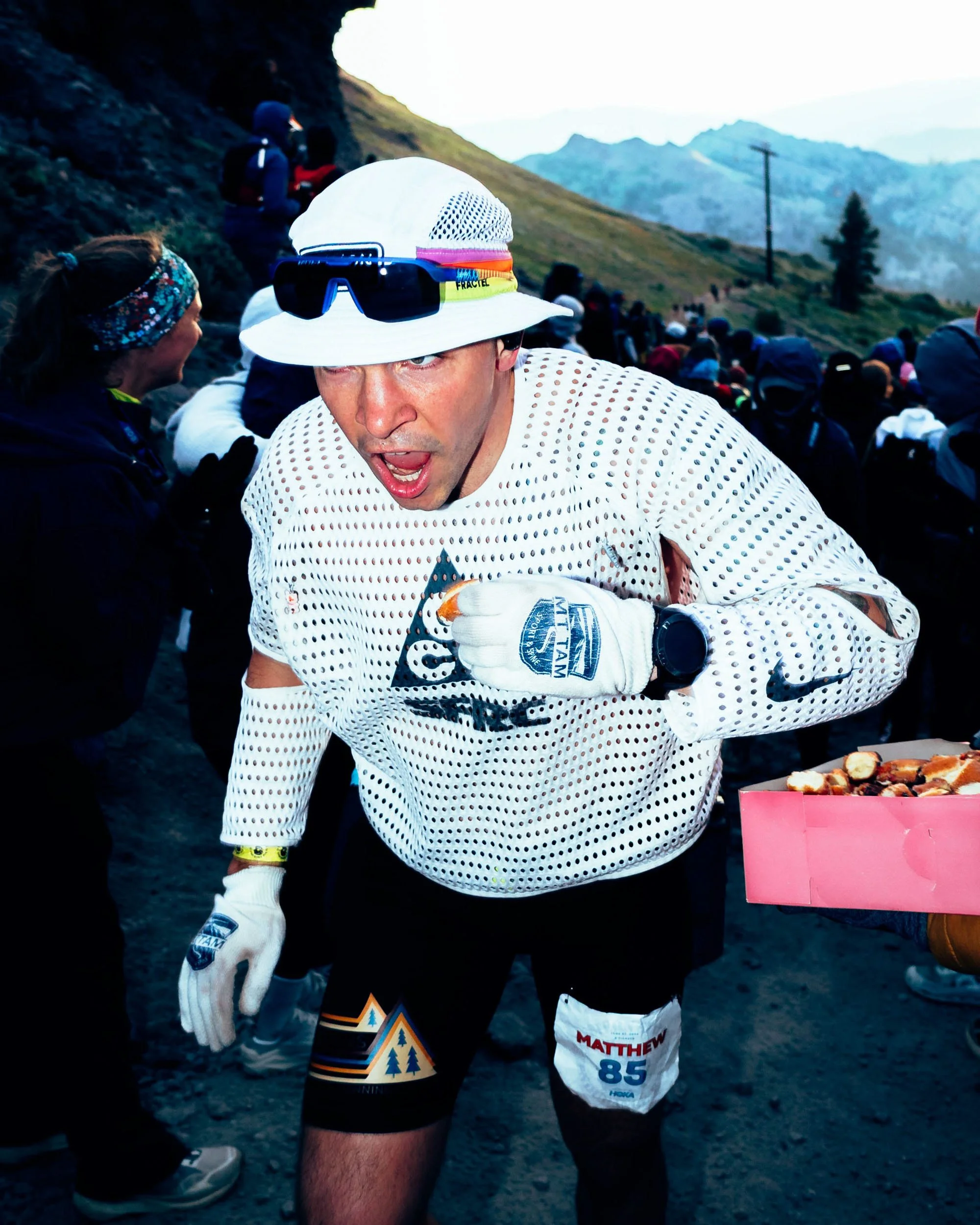 A man in a white mesh running shirt, black shorts with a race number, white gloves, and a white hat with sunglasses, participating in a trail race in a mountainous outdoor setting during daytime.