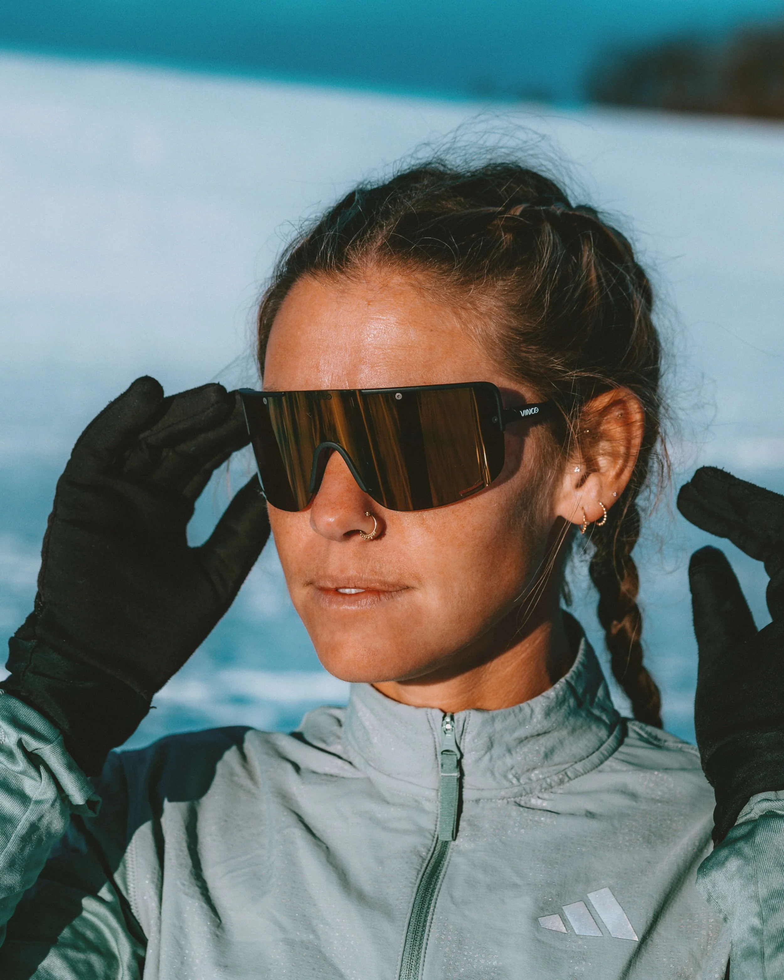 Close-up of a person with braided hair, wearing ski goggles, a light gray jacket, black gloves, and a backpack, outdoors in a snowy environment.