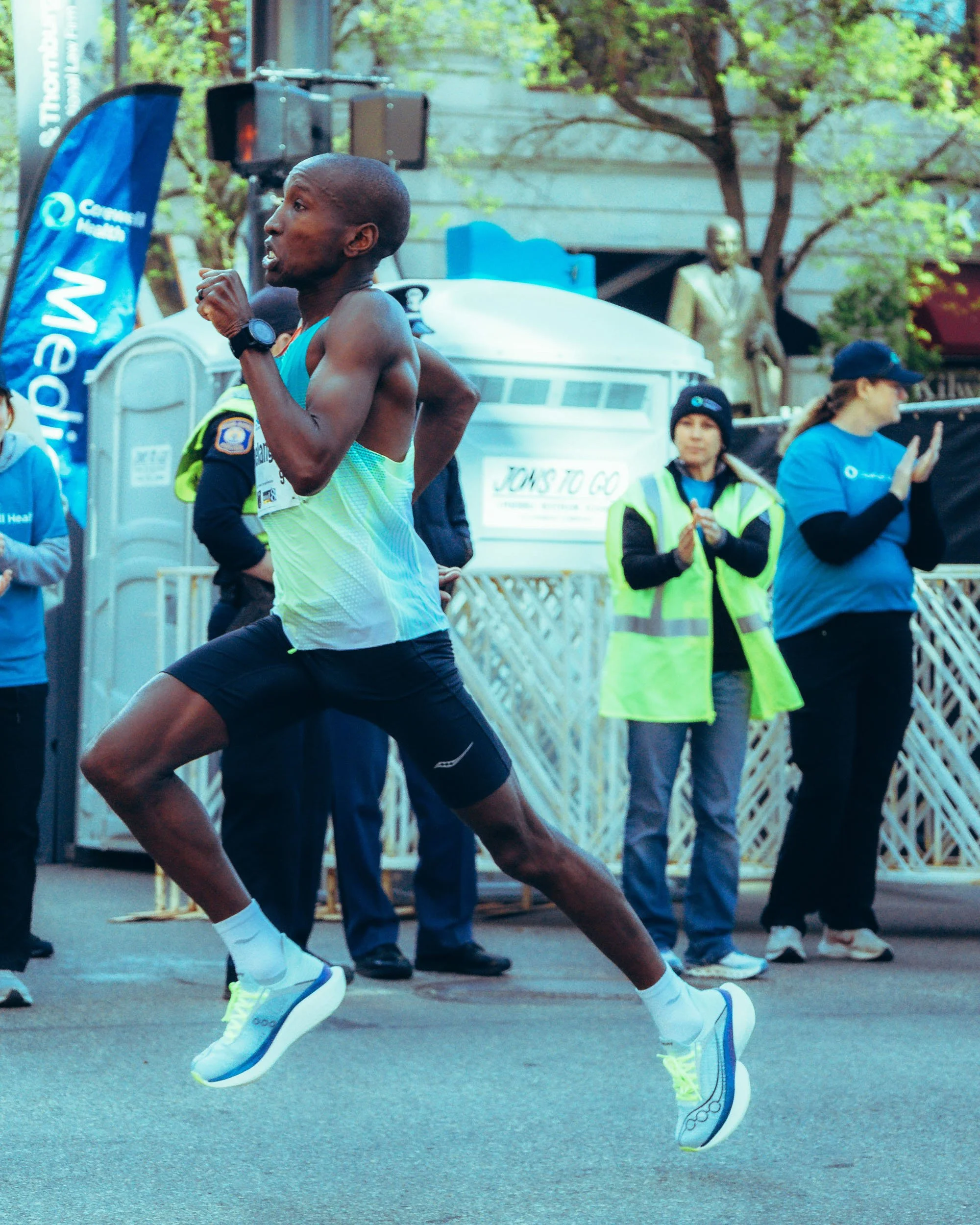 Close-up of a male marathon runner in motion, wearing a light blue sleeveless shirt, black shorts, white running shoes with neon yellow accents, and a watch on his wrist, running on a city street with spectators and trees in the background.