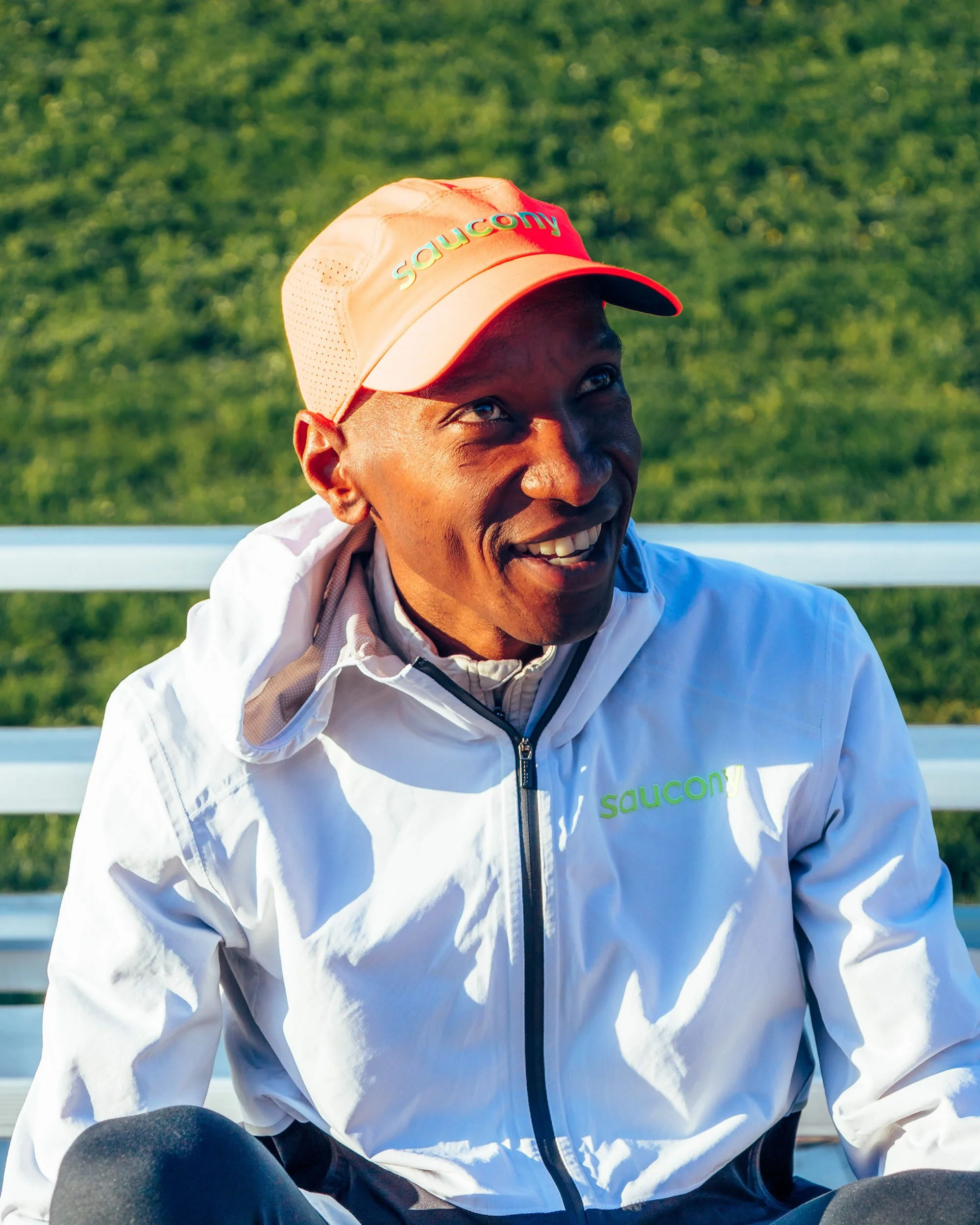 A person wearing an orange Saucony cap and a white jacket, smiling outdoors with green grass in the background.