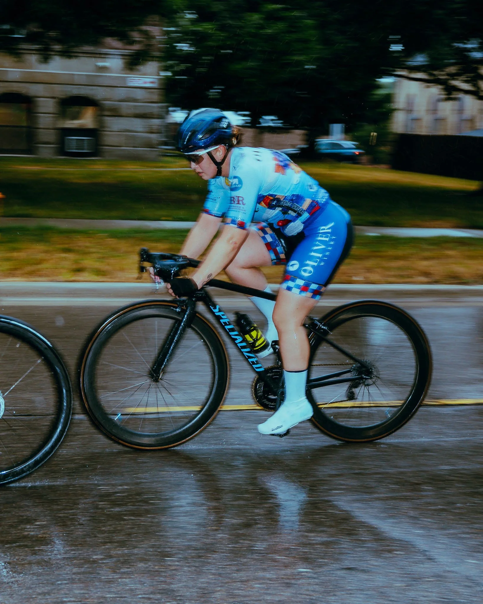A cyclist wearing a blue helmet and colorful cycling outfit riding a black Specialized road bike on a wet street, with motion blur indicating fast movement, and trees and buildings in the background.