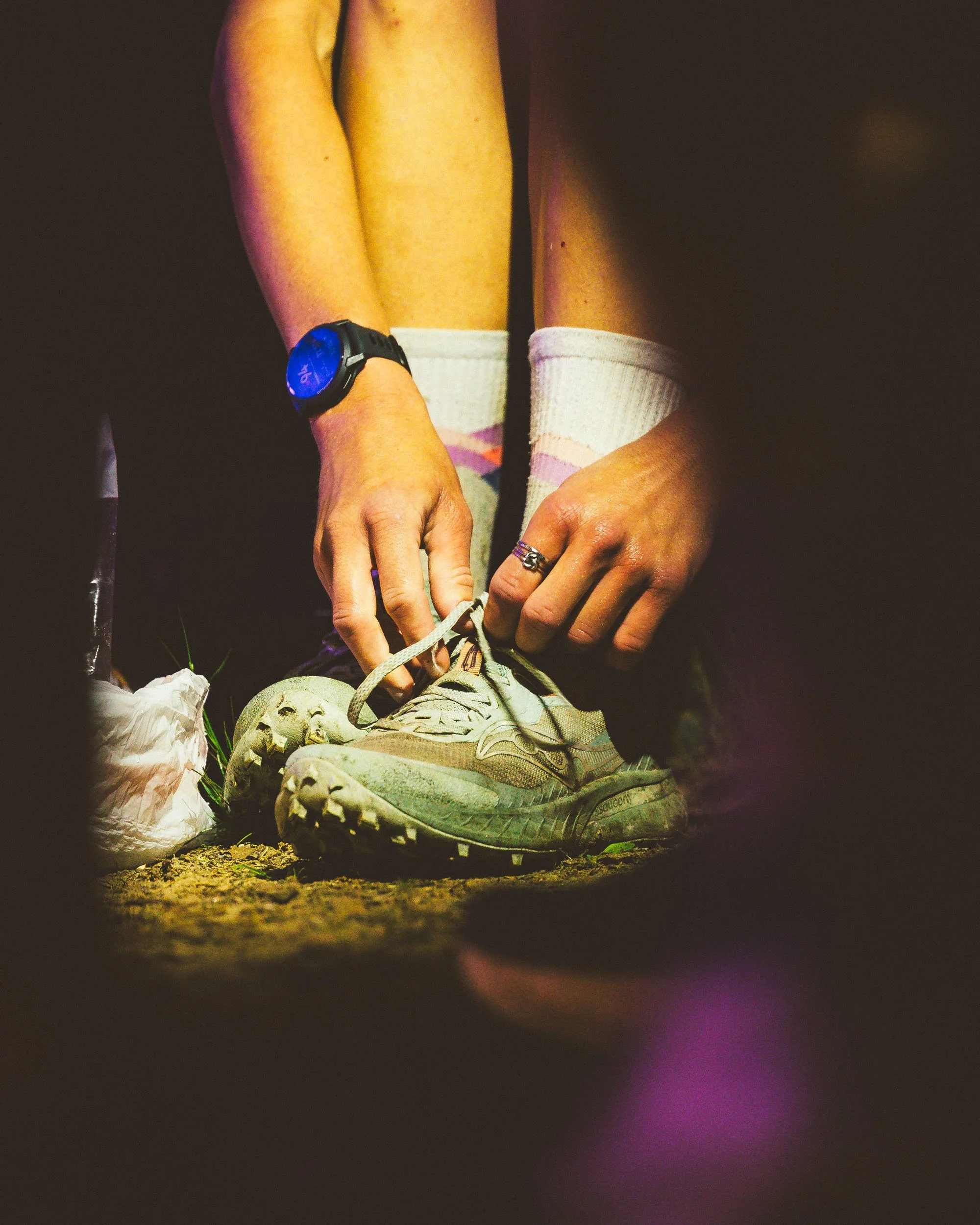 Close-up of a person tying shoelaces on a worn sneaker, wearing socks with pink and white stripes, with a digital watch on the left wrist, on dark ground with a woman's hand adorned with rings visible.
