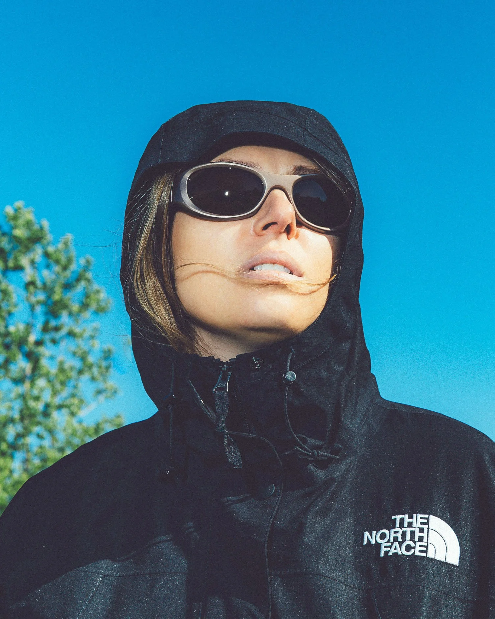 Close-up of a woman with long hair, wearing black sunglasses, a black hooded jacket, and a black The North Face jacket, standing outdoors against a bright blue sky with some trees visible.