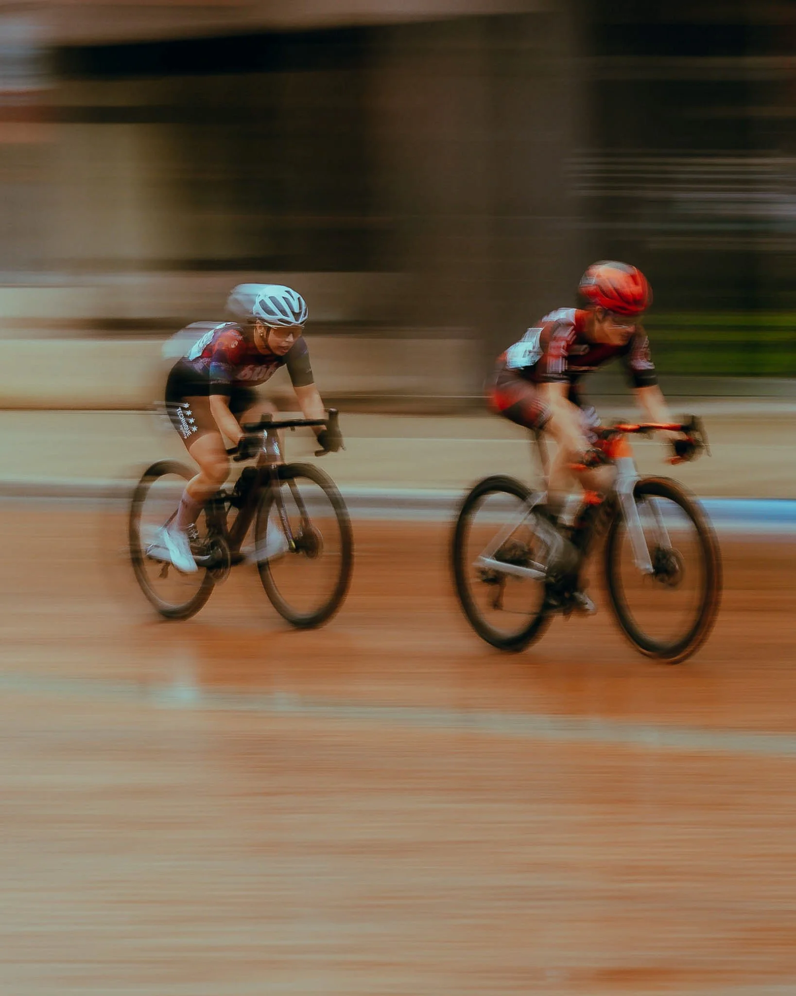 Two cyclists wearing helmets and sports gear riding fast on a dirt track, captured with motion blur.