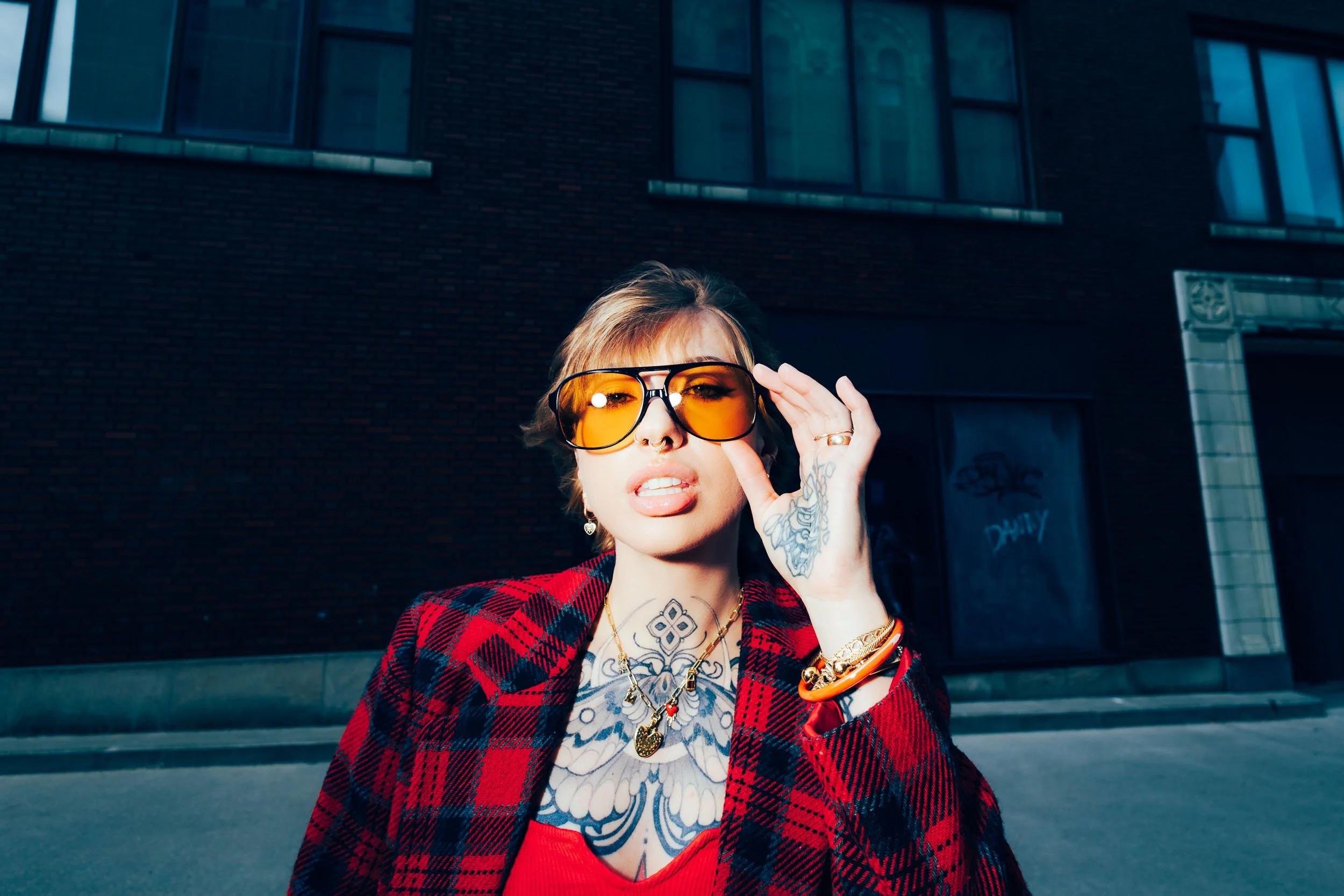 A woman with short hair and tattoos on her hand and neck adjusting her orange-tinted sunglasses outdoors against a dark brick building.