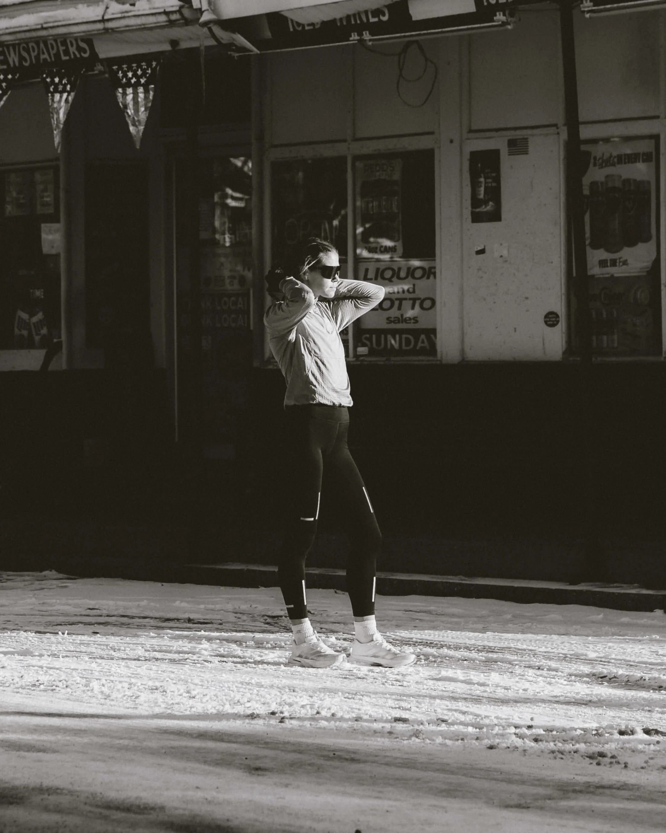 A woman wearing sunglasses, a jacket, and athletic shoes, stands on a snowy street with her hands behind her head. She appears to be in a town or city, with a storefront behind her displaying signs for liquor and lotto sales, and newspaper advertisem