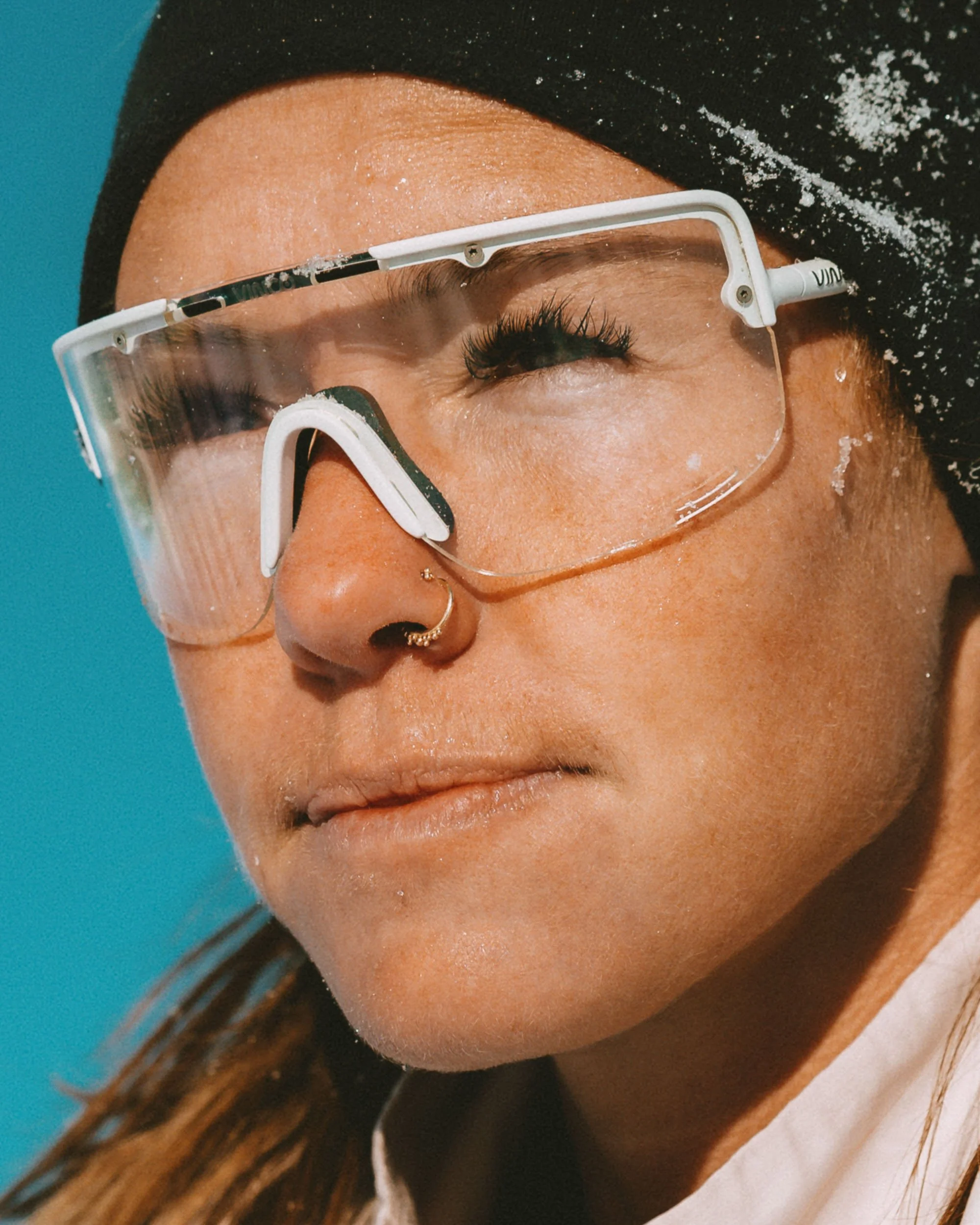Close-up of a woman wearing safety glasses, snow on her face, black beanie, and a white shirt, with a blue background.