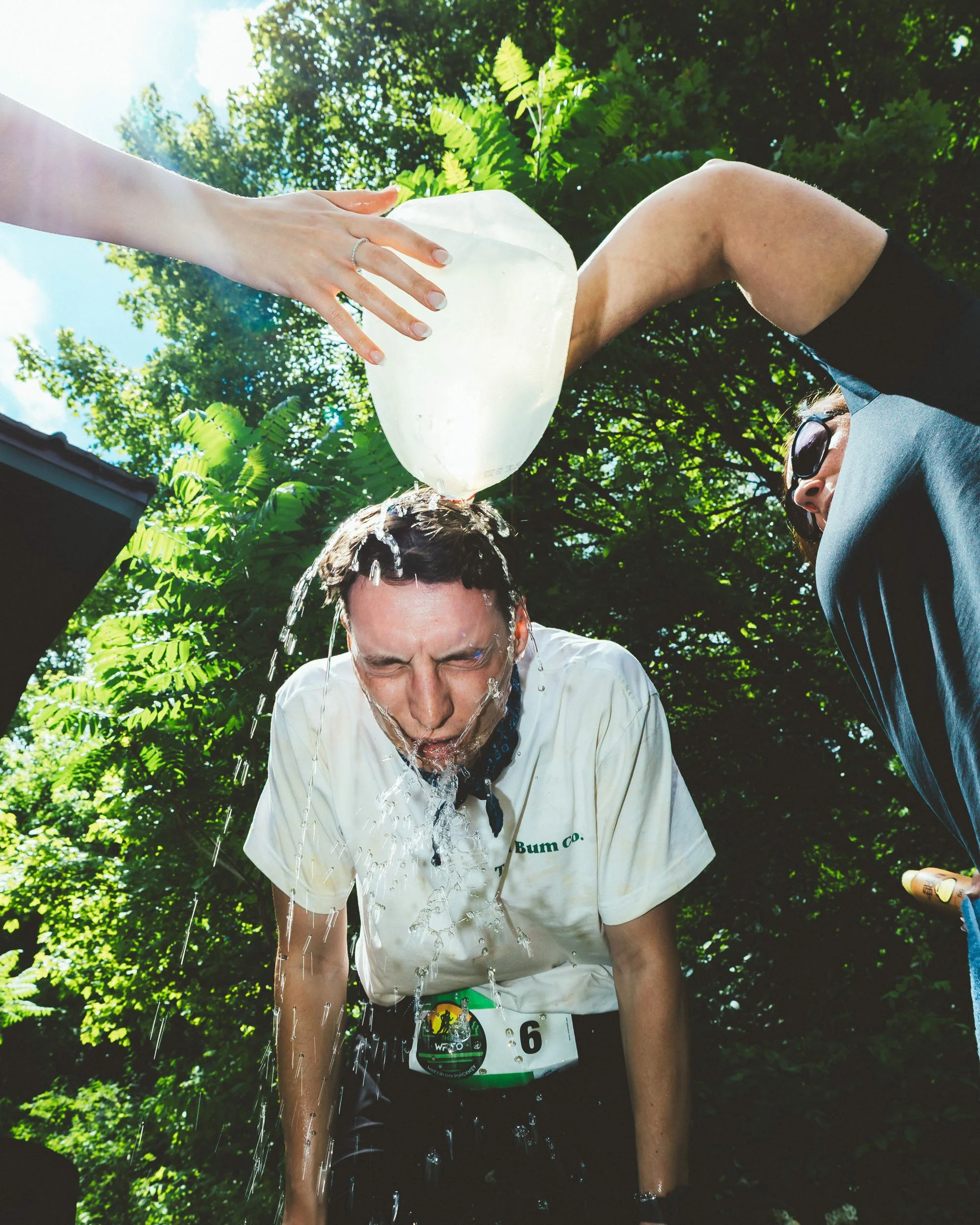 A man in a white shirt with a race number 6 is taking a shower outdoors. Another person is pouring water on his head from a large white container. There are lush green trees and bright sunlight in the background.