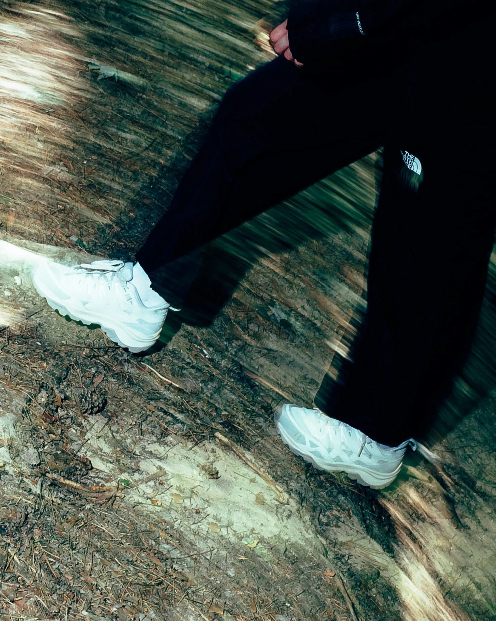 Close-up of a person's lower body with white sneakers and black pants, standing on a wooded forest floor with leaves and sticks.