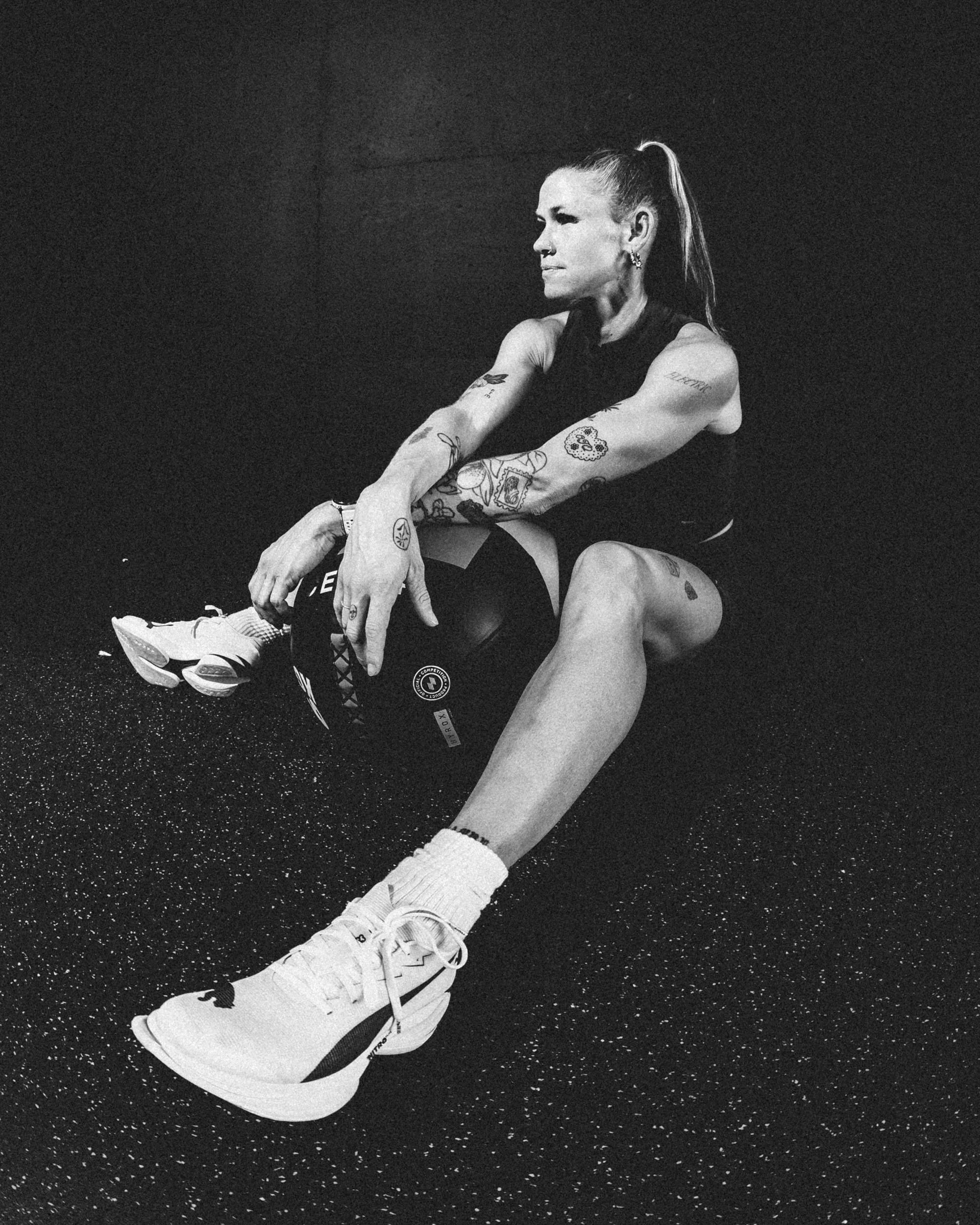 A woman with tattoos on arms and a ponytail, wearing sportswear, sits on a black speckled floor, tying her white athletic shoes.