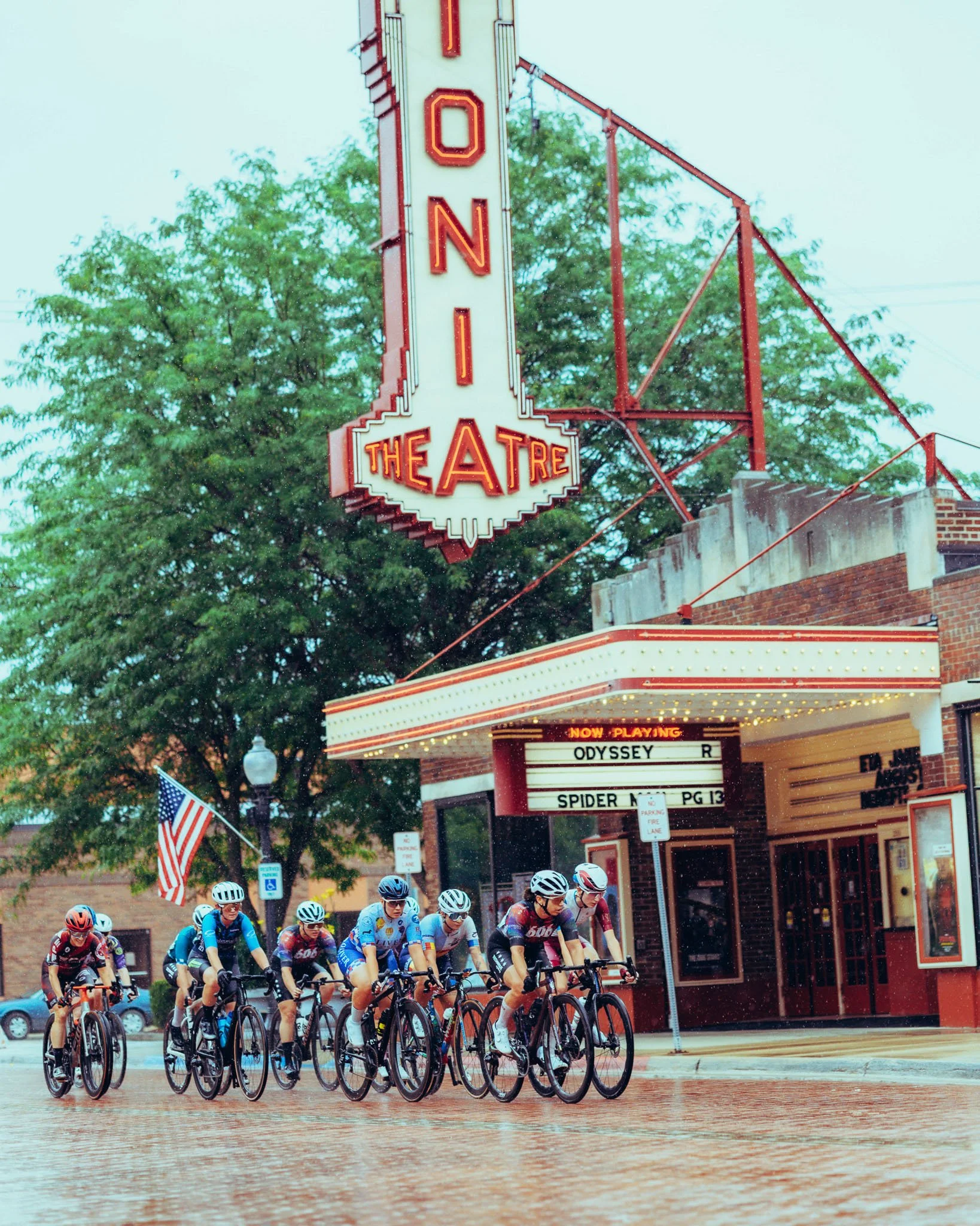 A group of cyclists riding in a race outside a theater with a marquee and neon signs, on a wet pavement during daytime.