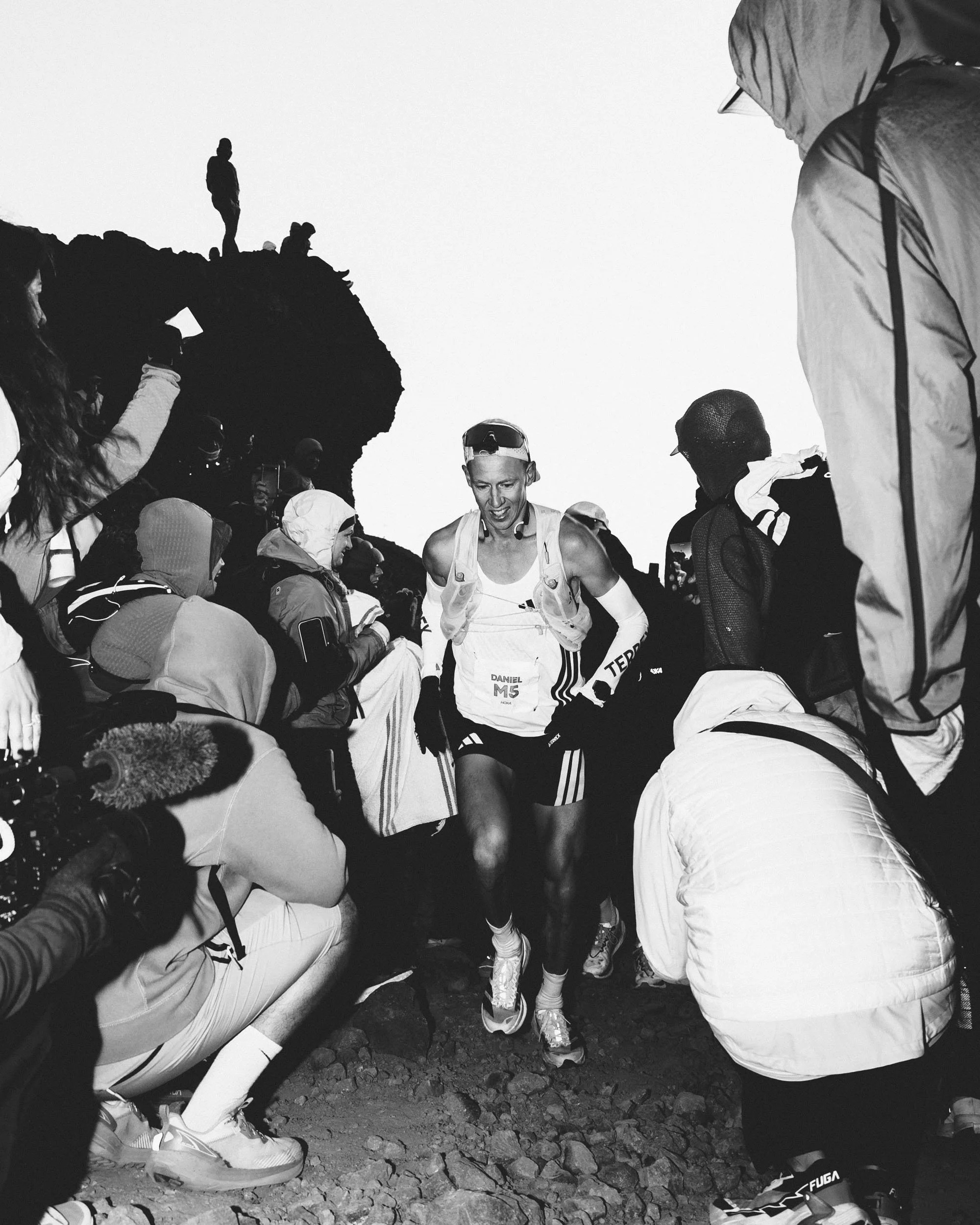 A group of hikers and a runner gathered outdoors on rocky terrain, some wearing hiking gear and jackets, with a silhouette of another person standing on a mountain in the background.