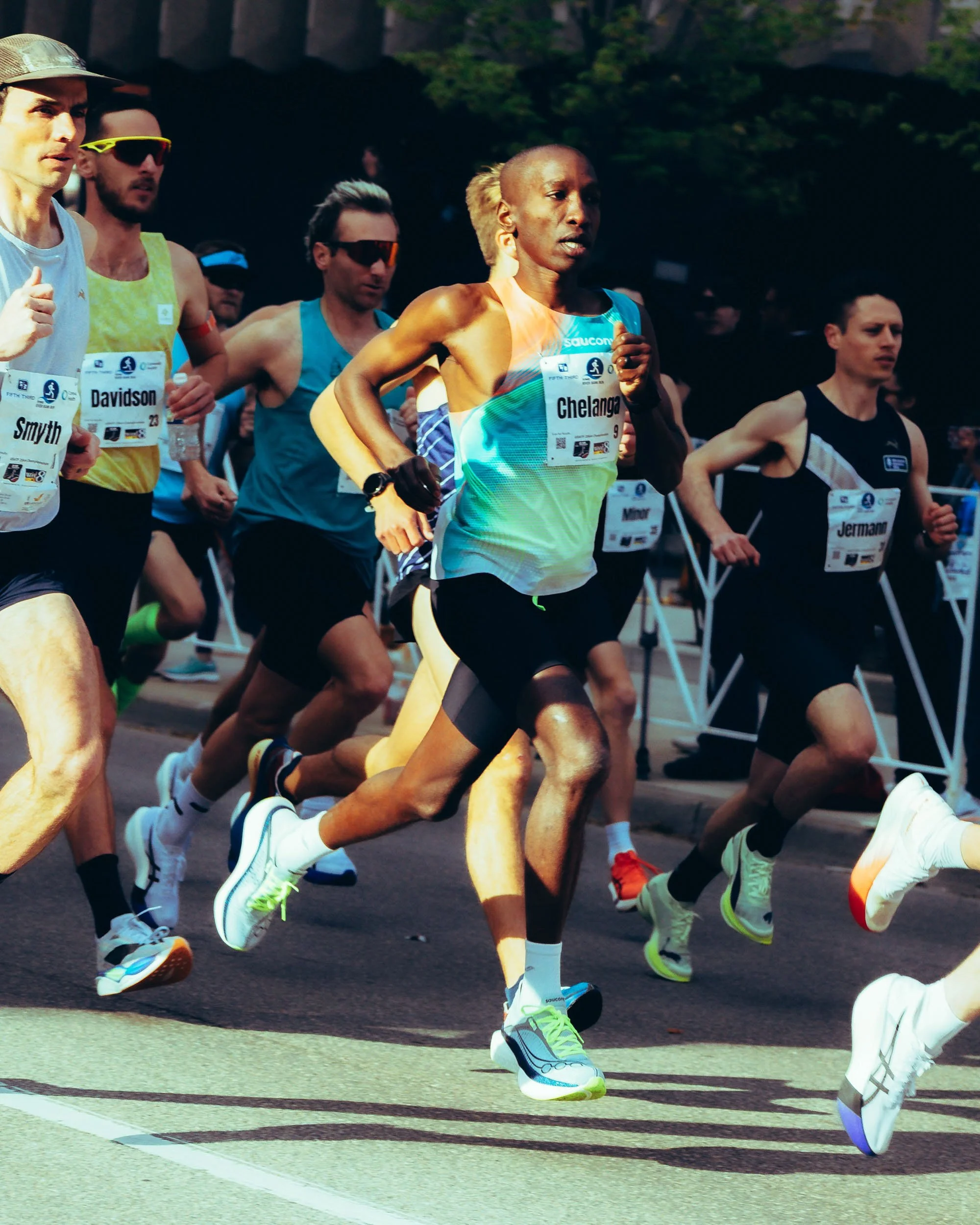 Group of runners participating in a race, wearing athletic gear and race bibs, running on a street.