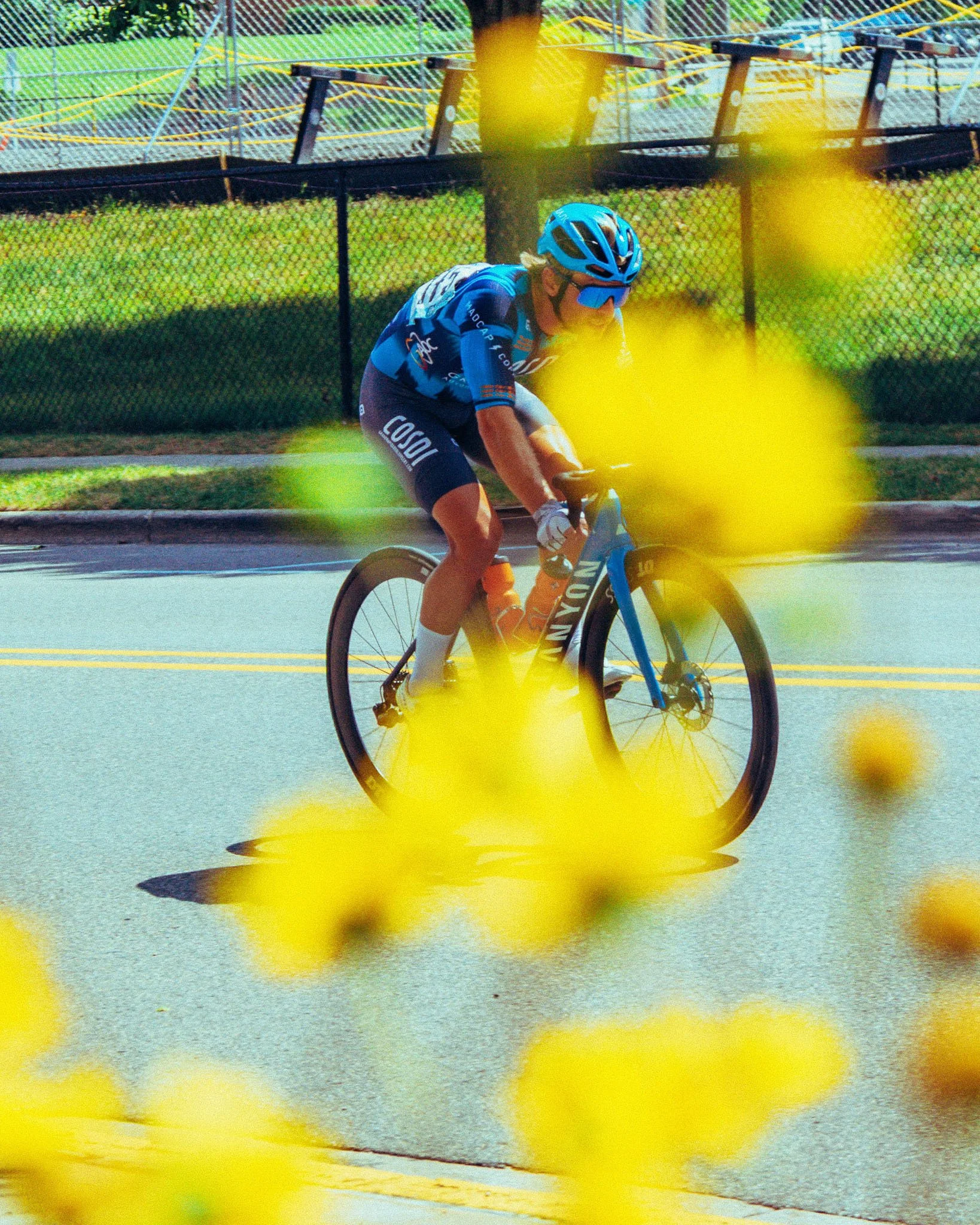 A cyclist wearing a blue helmet and matching blue cycling jersey riding a bicycle on a street, with yellow flowers in the foreground, a black fence, and stairs in the background.