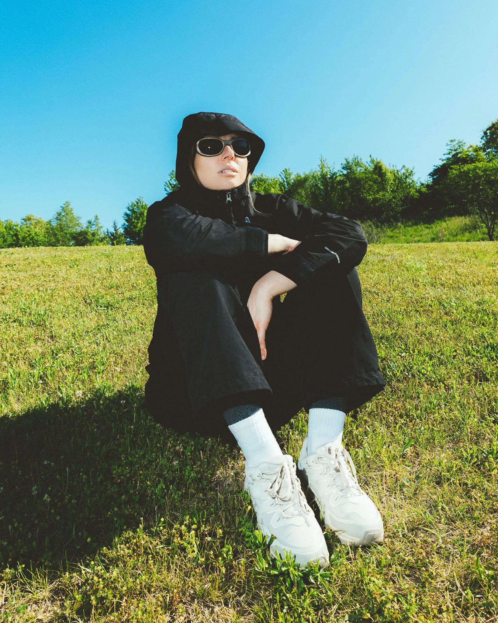 Person wearing a black hooded jacket, sunglasses, and black pants sitting on green grass in an open field, with trees and a clear blue sky in the background.