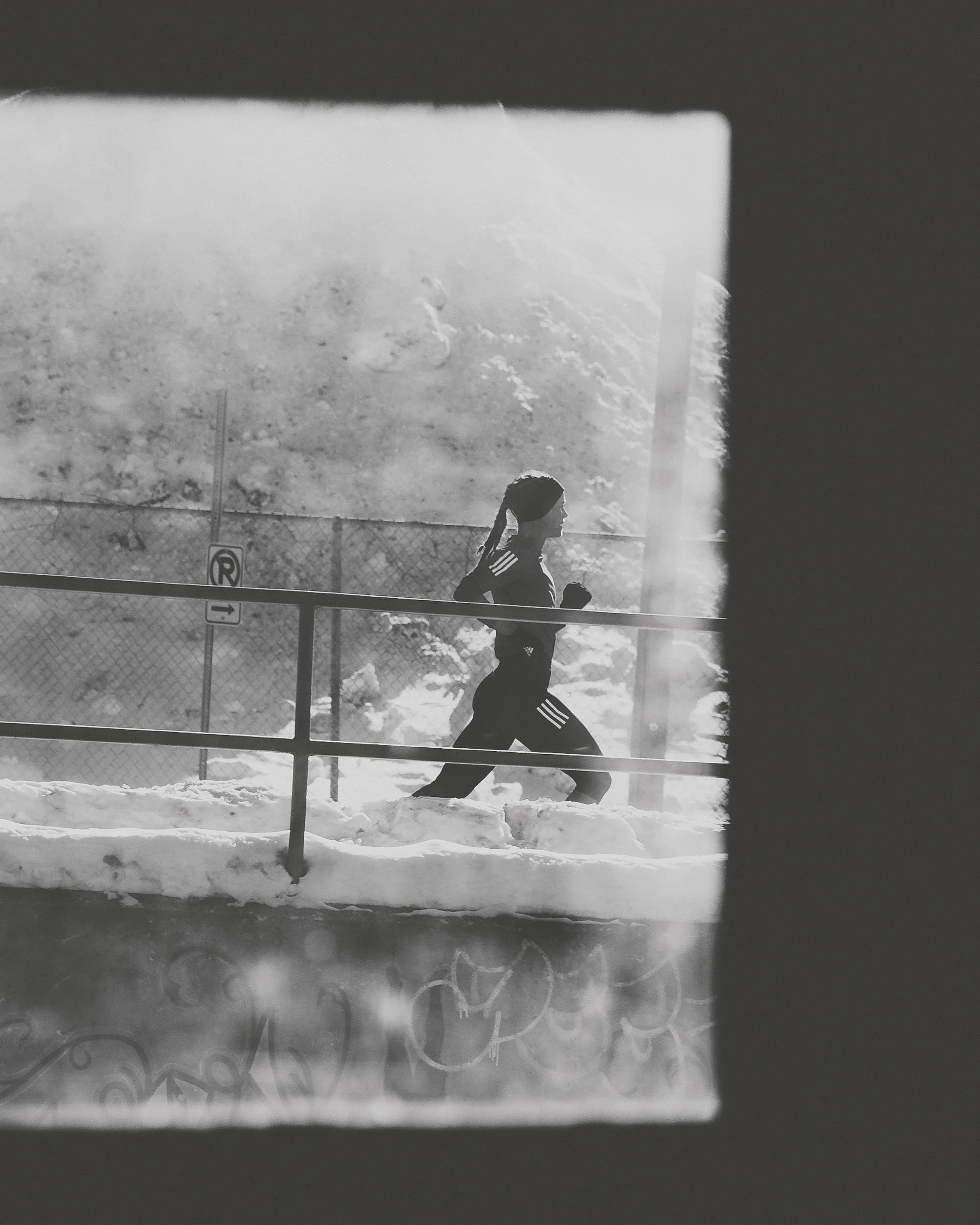 A person jogging outside in the snow, viewed through a window with frosted and clear sections, including a fence and signs in the background.